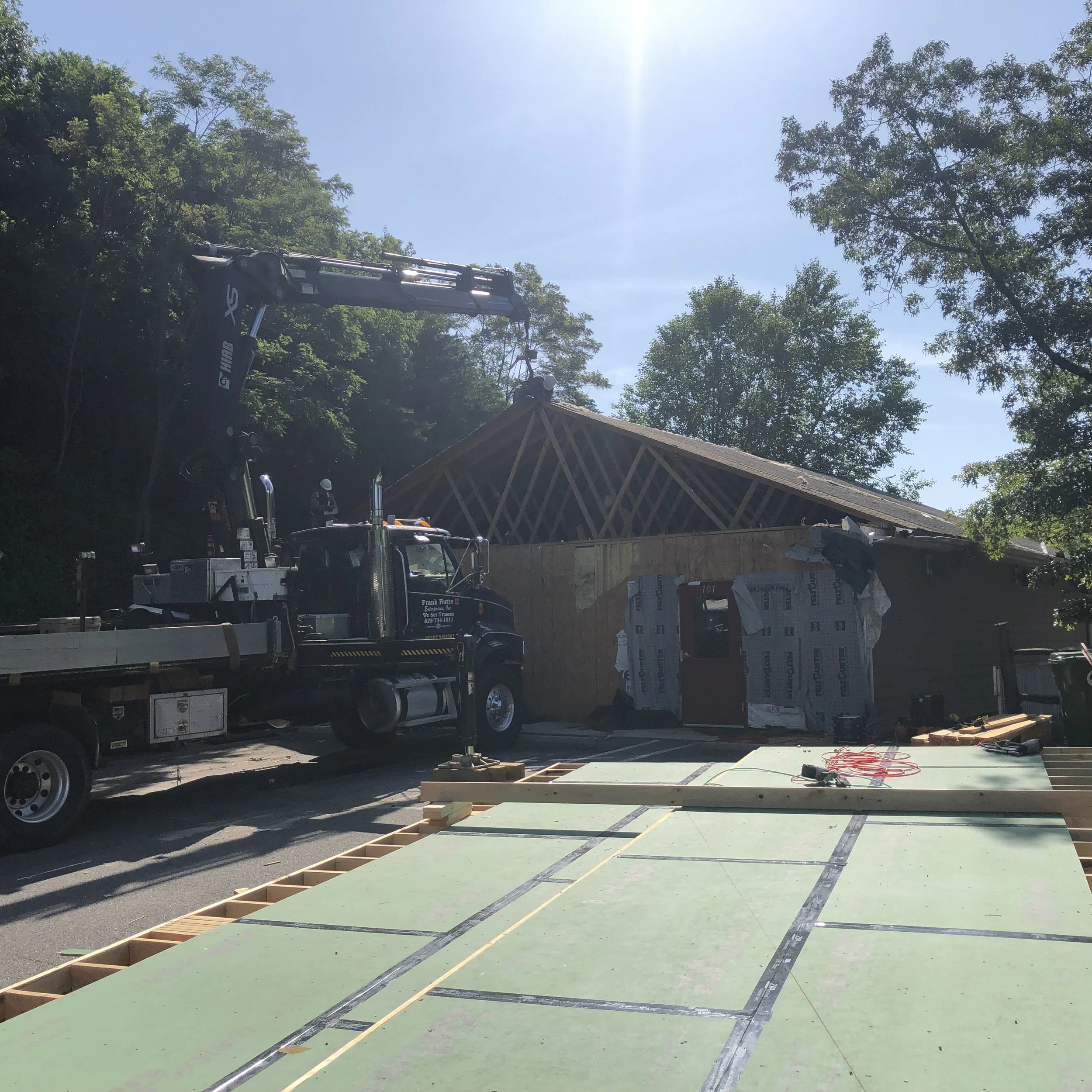 A construction site with a house under renovation, a crane lifting a roof section, and building materials on the ground.