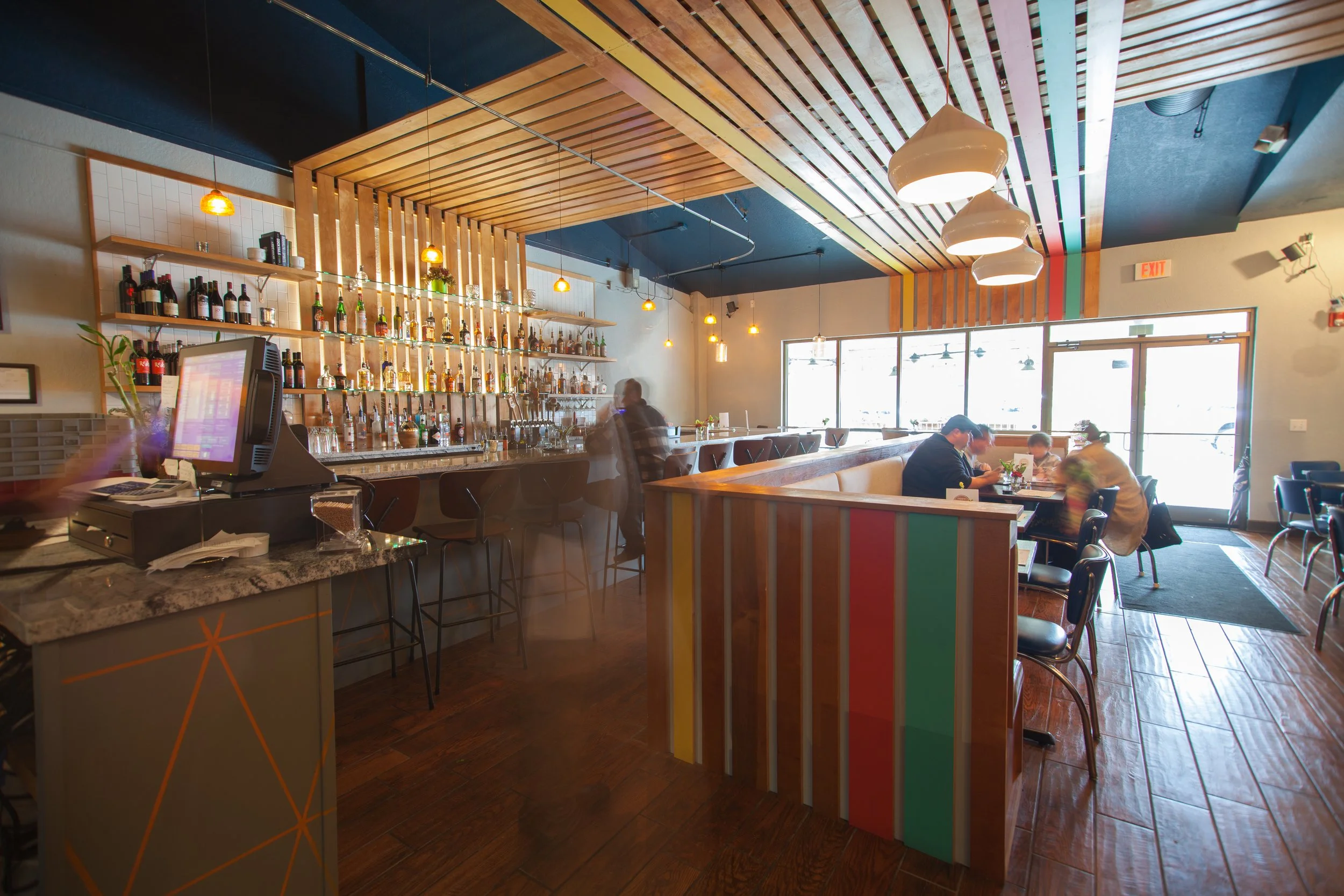Interior of a modern restaurant or cafe with a bar area, shelves of liquor bottles, hanging lights, and customers seated at tables near large windows.