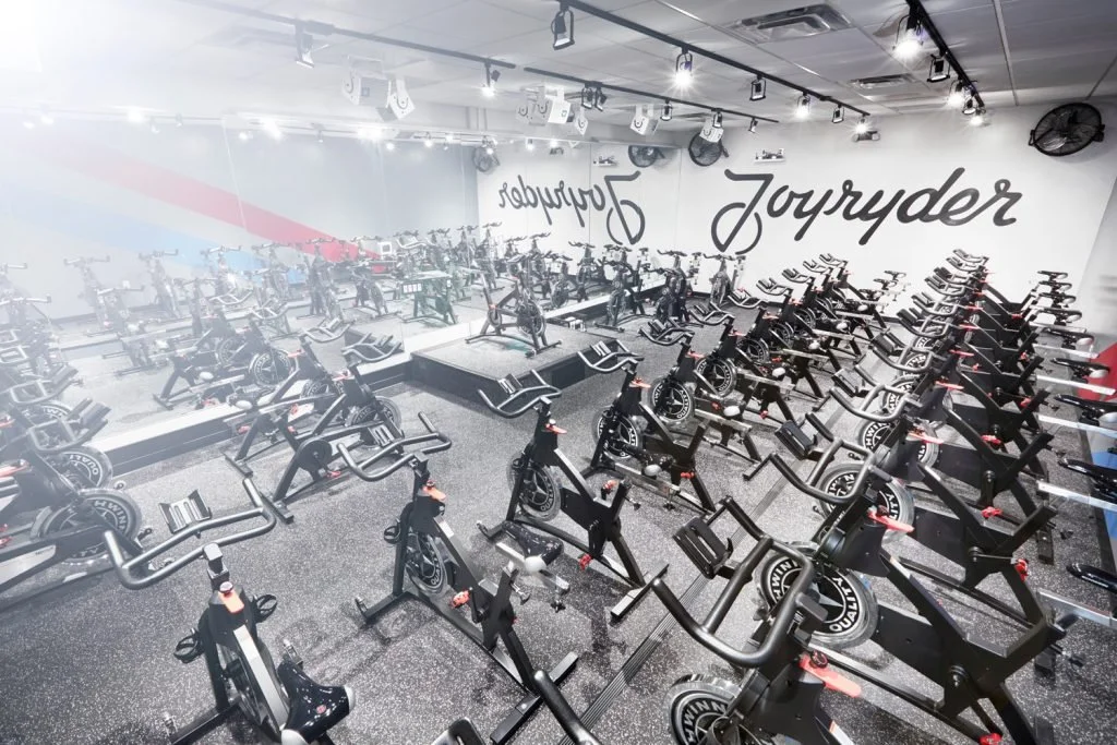 Indoor gym filled with multiple stationary cycling bikes and a raised platform at the front, with words "Joyrider" and "Prowler" on the wall.
