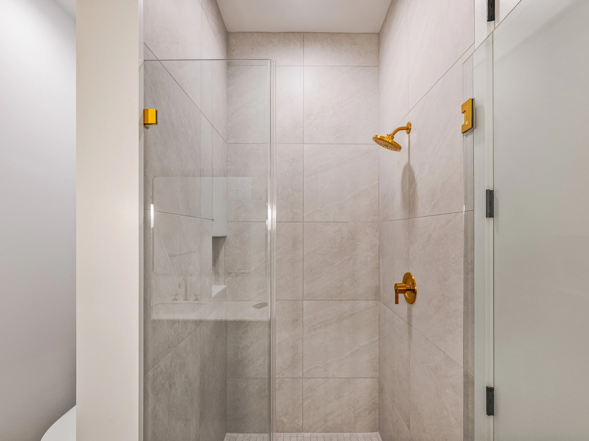 Modern walk-in shower with beige tiles, a glass door, a gold showerhead, and a small built-in shelf.