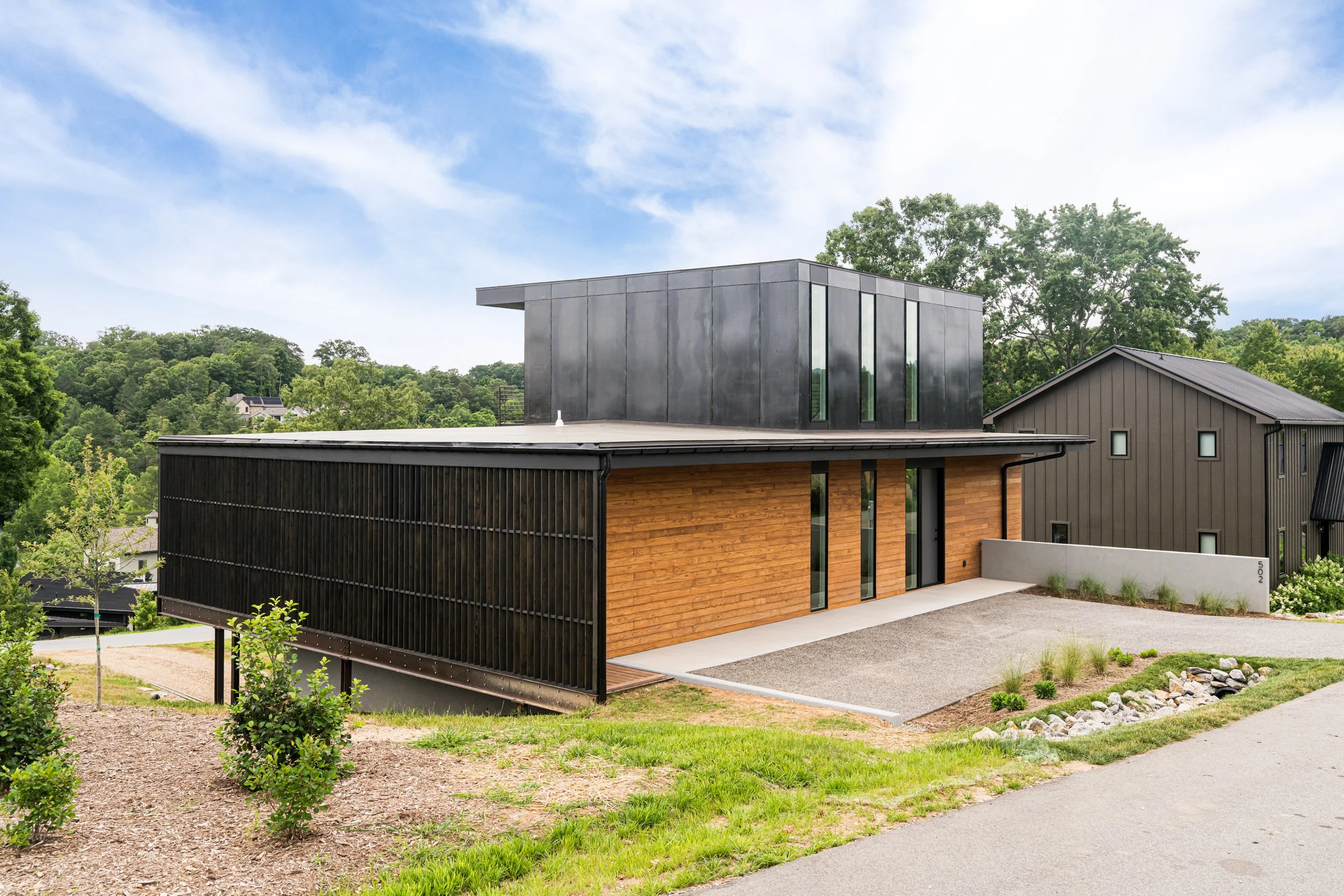 Modern house with black metal and wooden exterior, surrounded by green landscape and trees, under a partly cloudy sky.