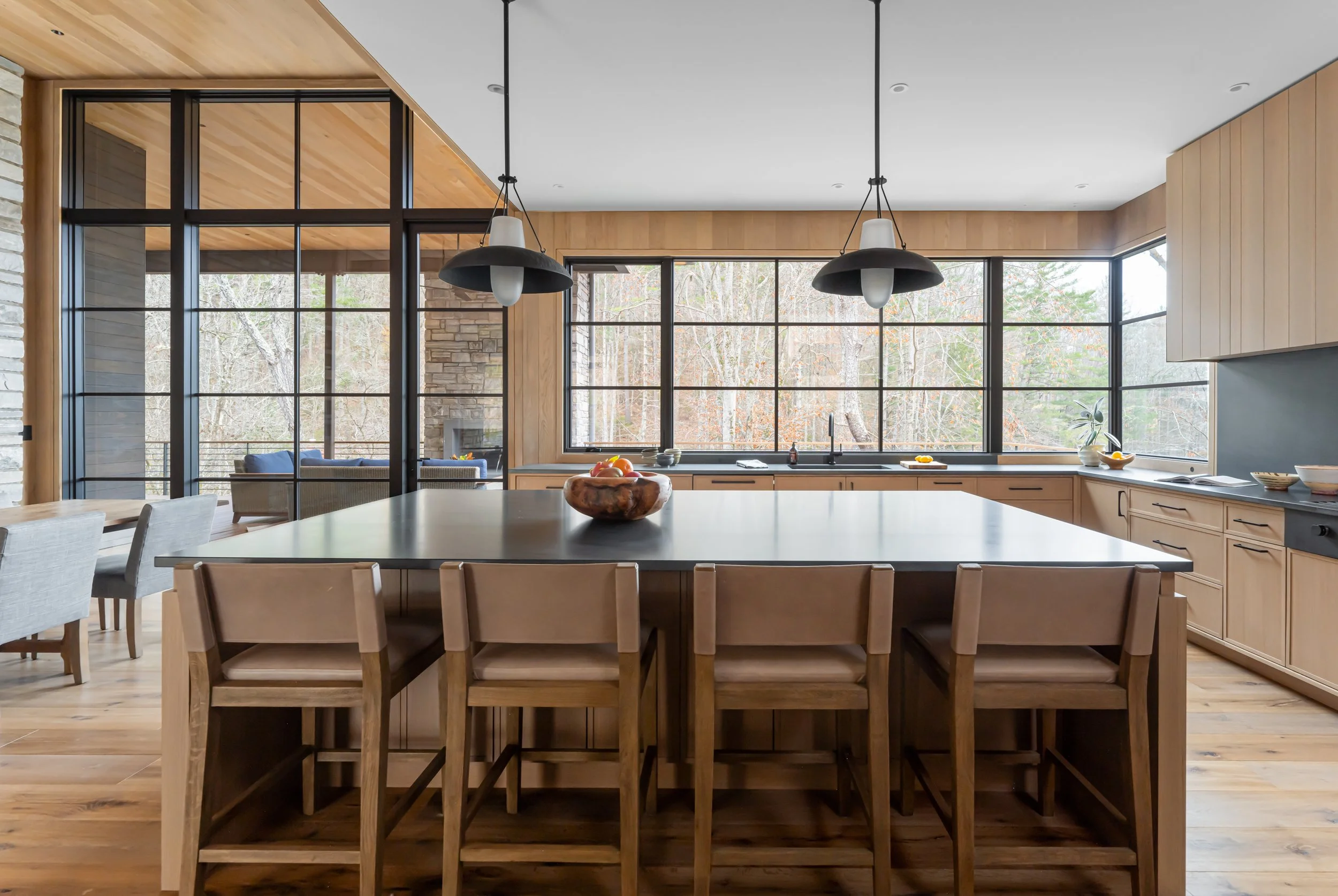 Modern kitchen with large central island, wooden cabinetry, black pendant lights, and expansive windows overlooking trees.