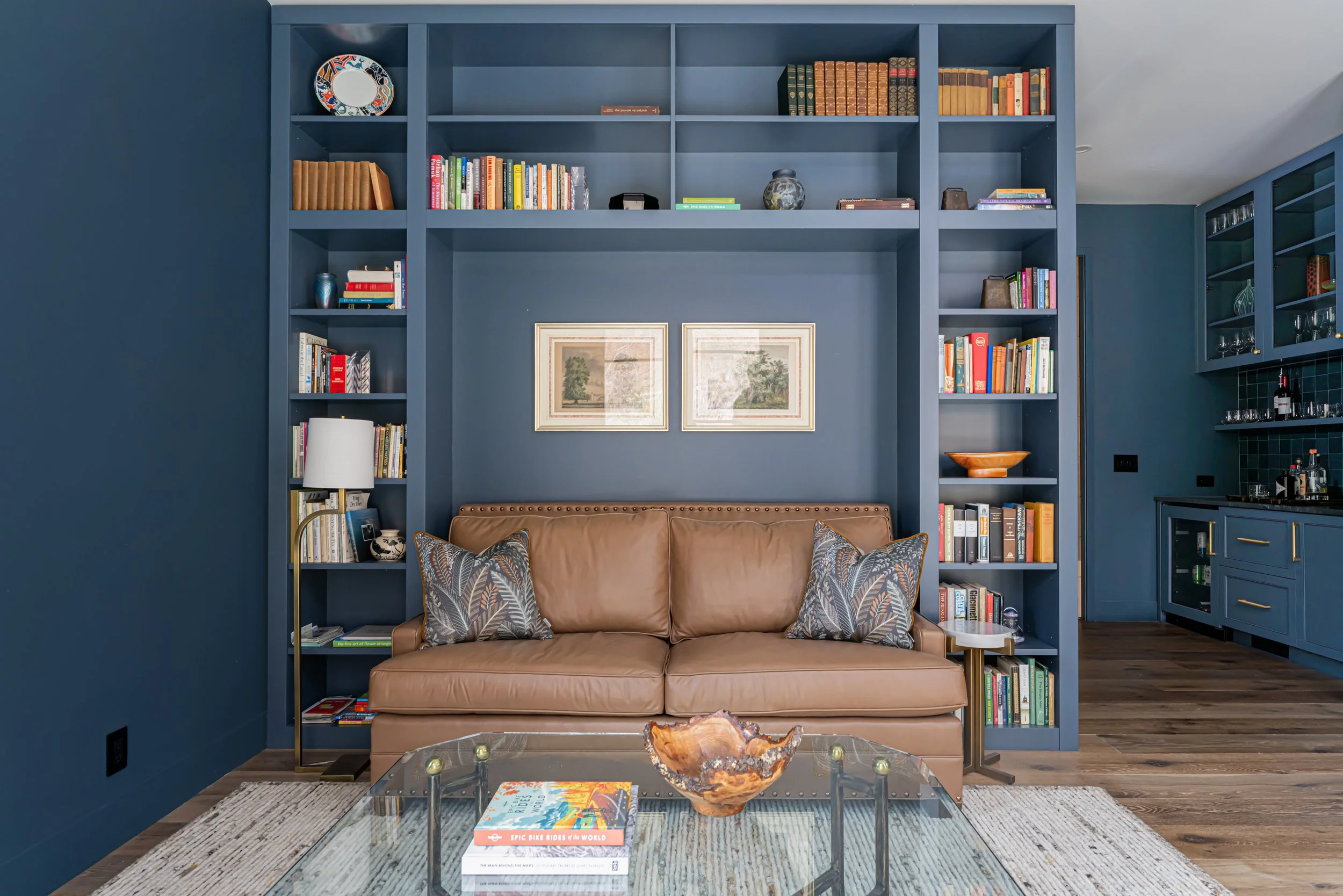 Living room with a brown leather sofa, decorative pillows, glass coffee table with a wooden bowl and books, navy blue built-in bookshelf, framed artwork on the wall, wood flooring, and a navy blue kitchen area to the right.