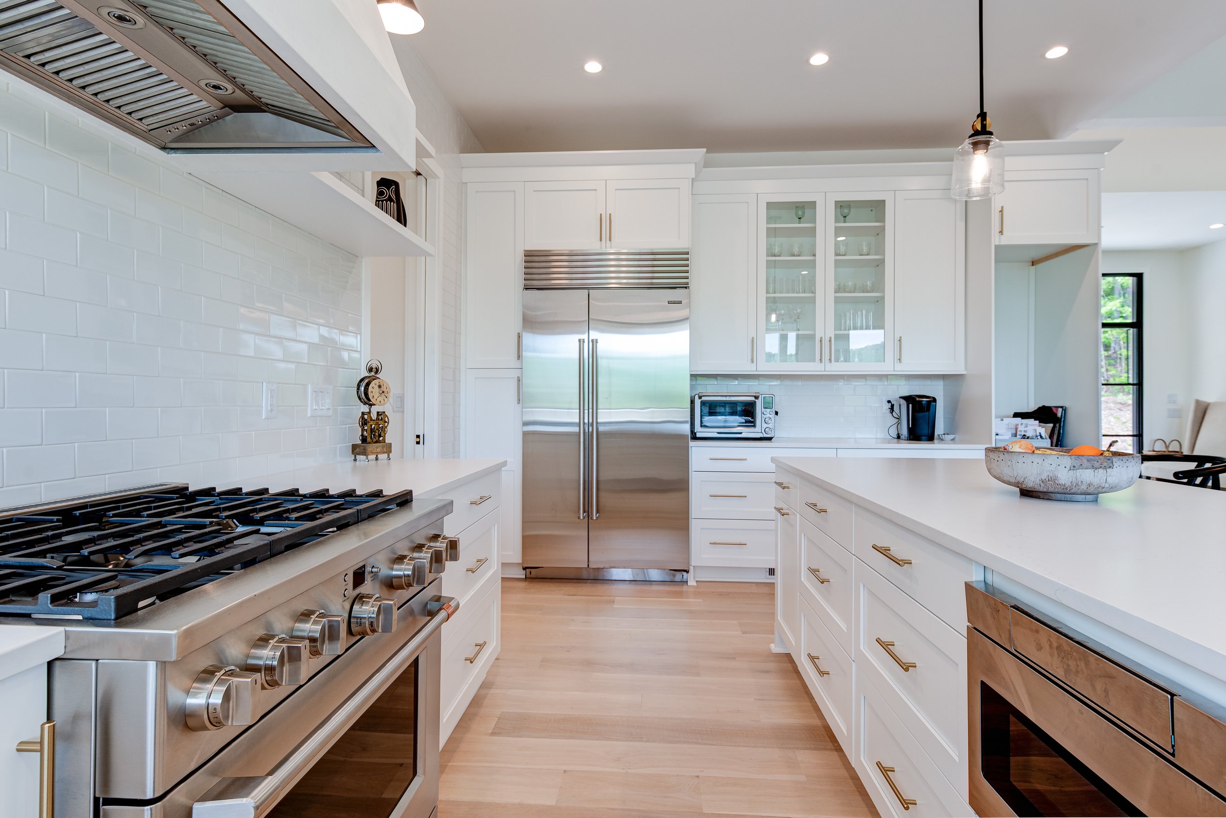 Modern white kitchen with stainless steel refrigerator, oven, microwave, coffee maker, and pendant light, with wooden flooring and cabinets with gold handles.