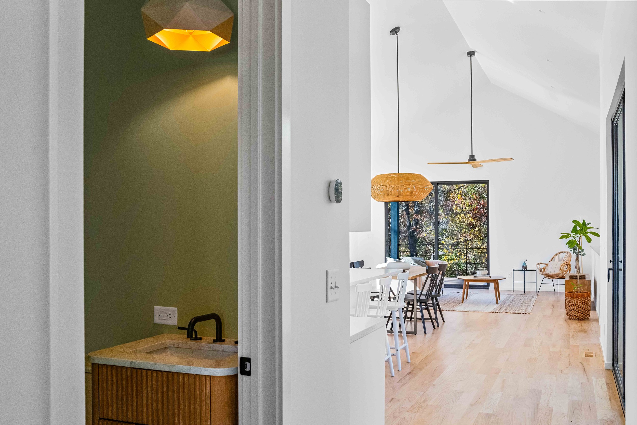 A spacious, modern living and dining area with large windows, wooden floors, and minimalist furniture. There are two hanging pendant lights, a ceiling fan, a potted plant, and a cozy chair with a small side table.