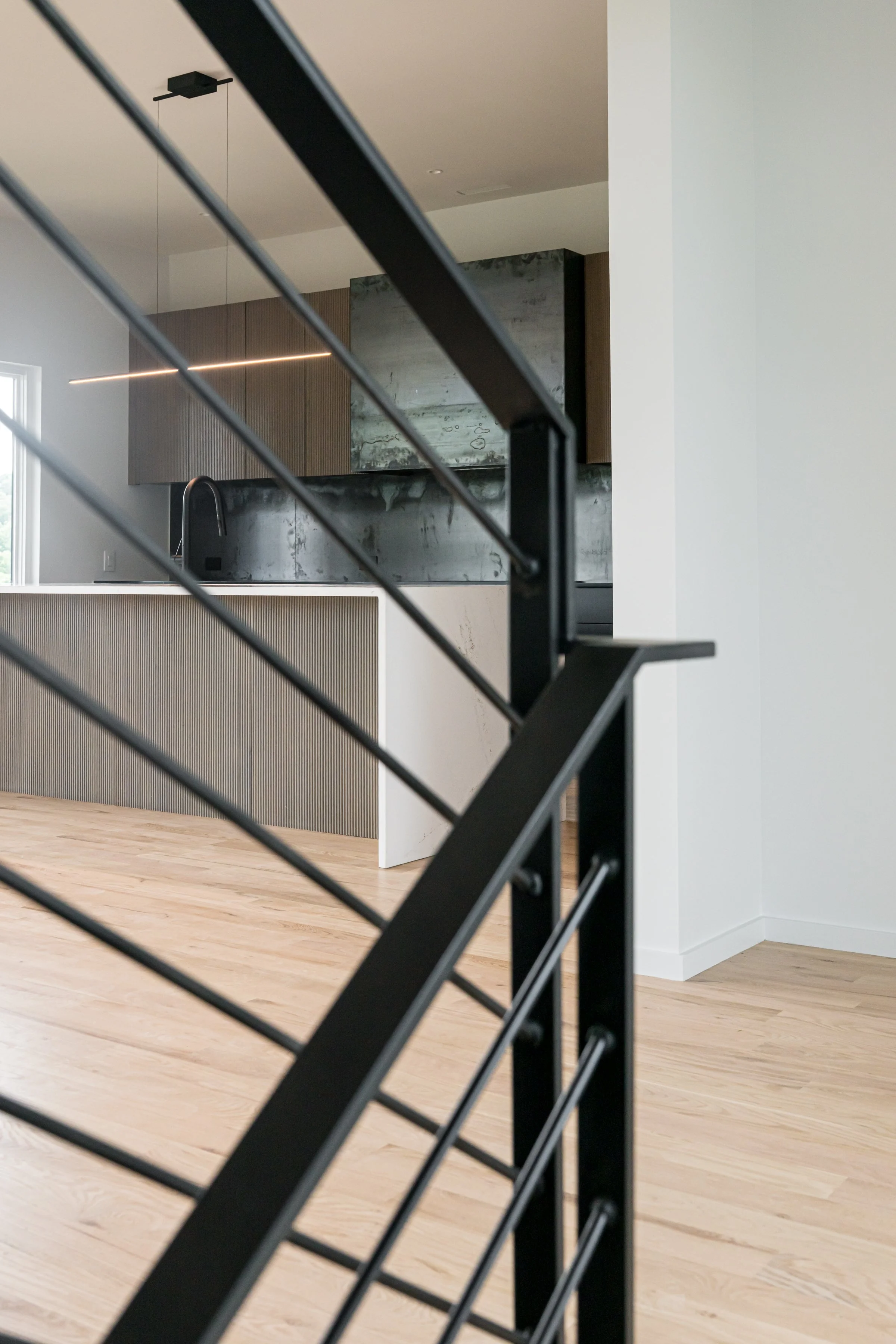 Modern kitchen with black metal railing, light wooden floors, and minimalist cabinetry and countertops.