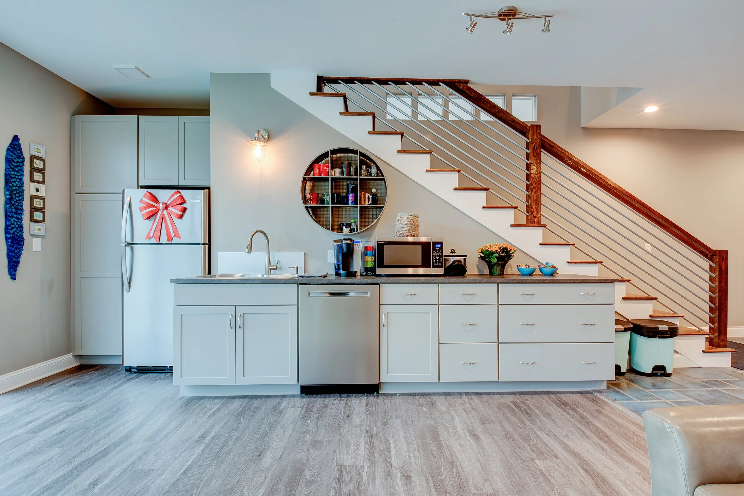 Kitchen area under stairs with white cabinets, a microwave, a refrigerator with a red bow decoration, a round wall shelf with mugs, and a staircase with wooden railing and metal spindles.