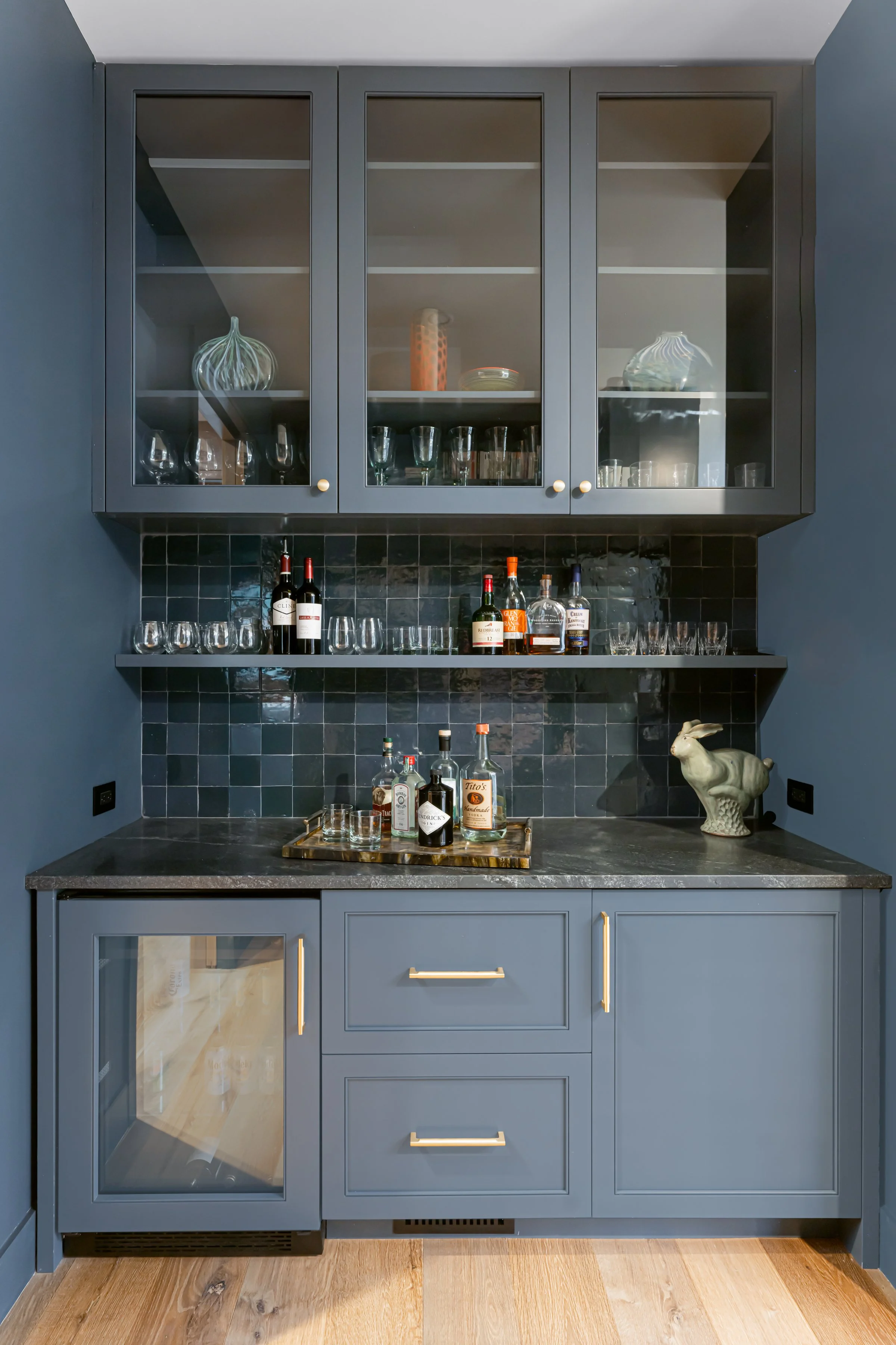 A home bar with dark blue cabinetry, a black tiled backsplash, and a black countertop. The upper cabinets have glass doors and display glassware and decorative items. A shelf holds liquor bottles and glassware. The lower cabinet has a small wine frid