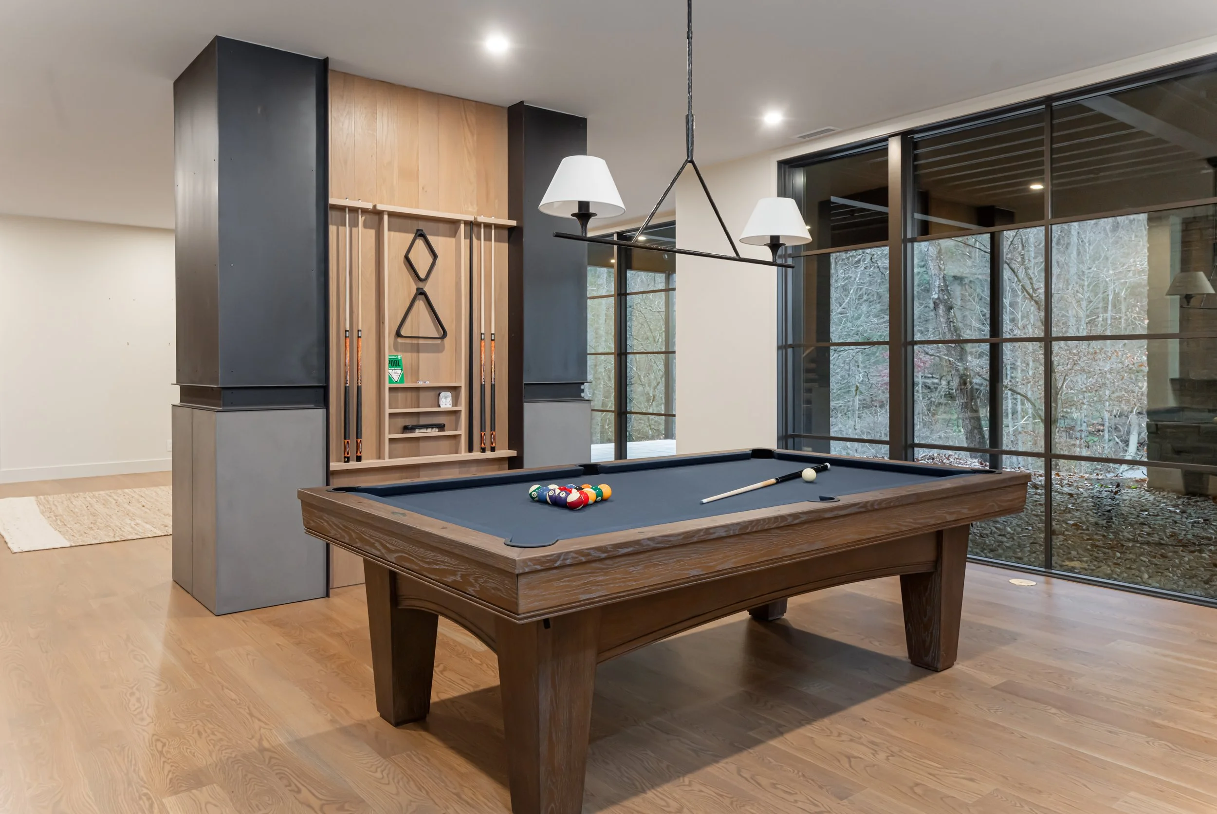 Indoor game room with a pool table, pool cues, and a rack against the wall. Large windows reveal an outdoor view of trees and fallen leaves.