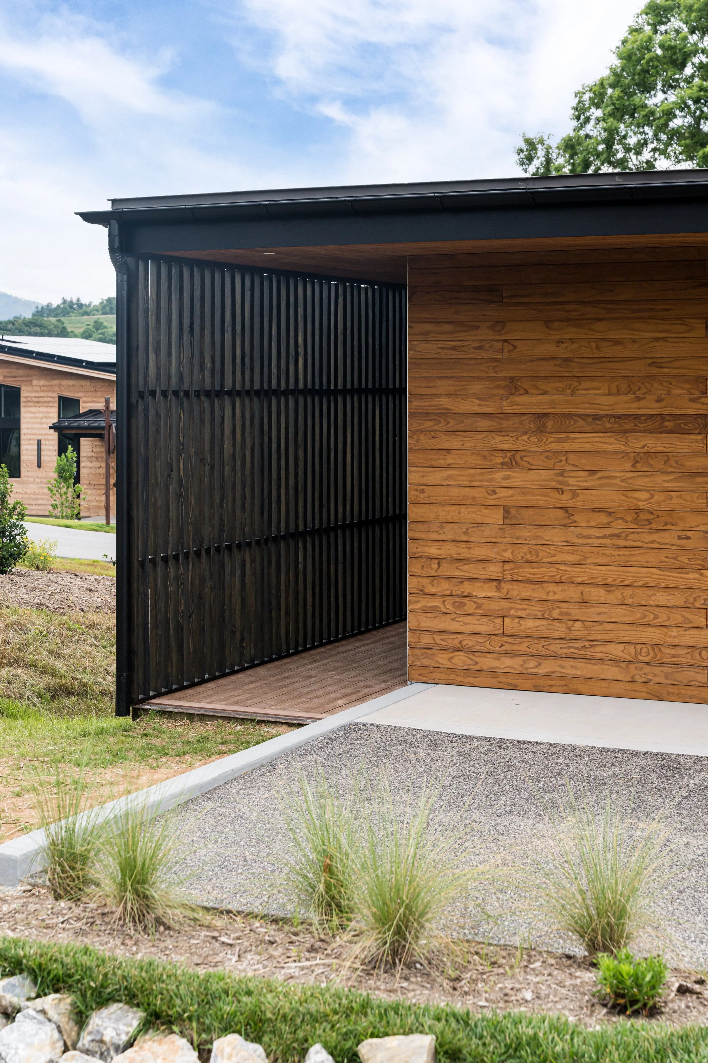 Modern house exterior with black vertical slat wall, wood panel wall, concrete and gravel pathway, and landscaping with grasses and rocks.
