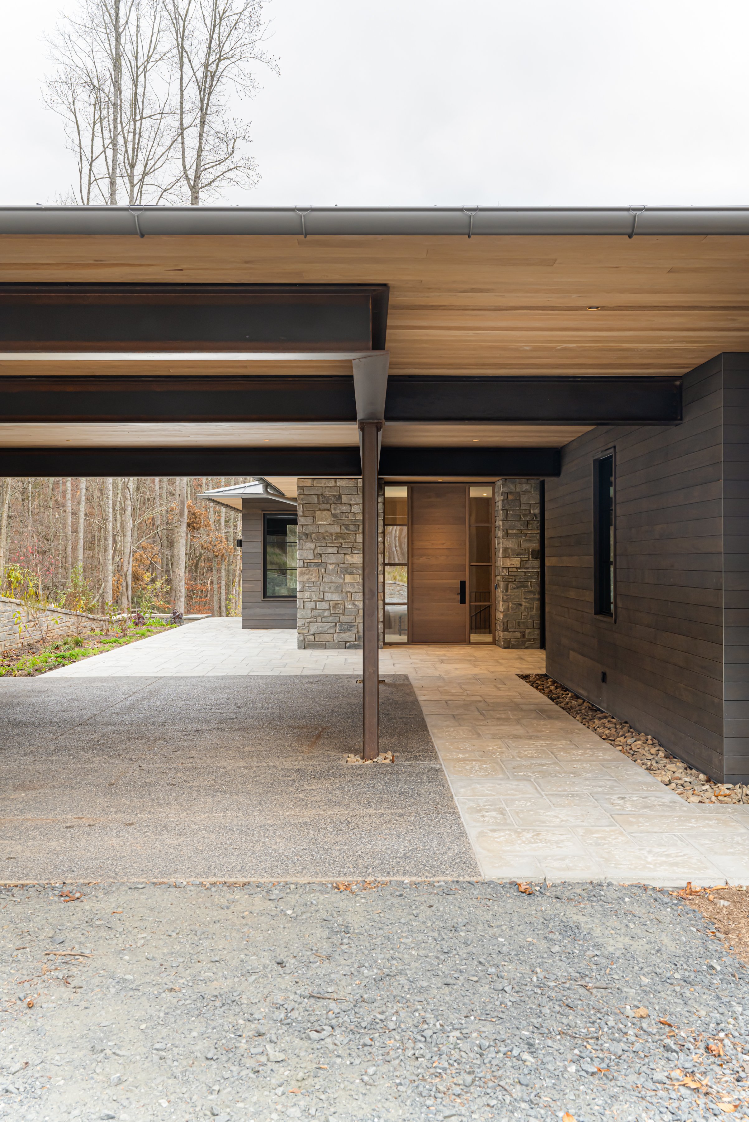 Modern house exterior with a drive-through carport, stone and dark wood siding, and a paved walkway leading to the front door, surrounded by trees.