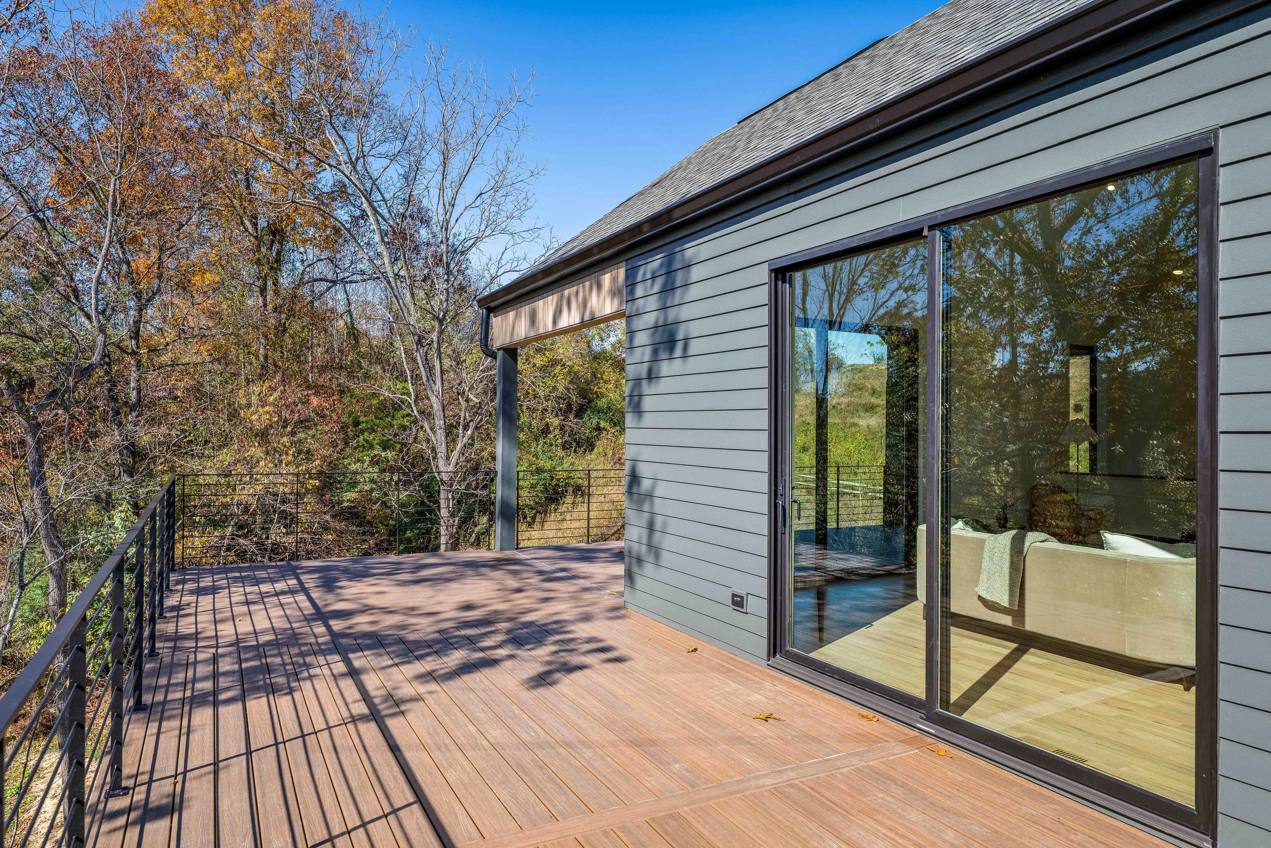 A spacious wooden deck attached to a modern house with large glass sliding doors, overlooking a wooded area with trees in fall foliage.