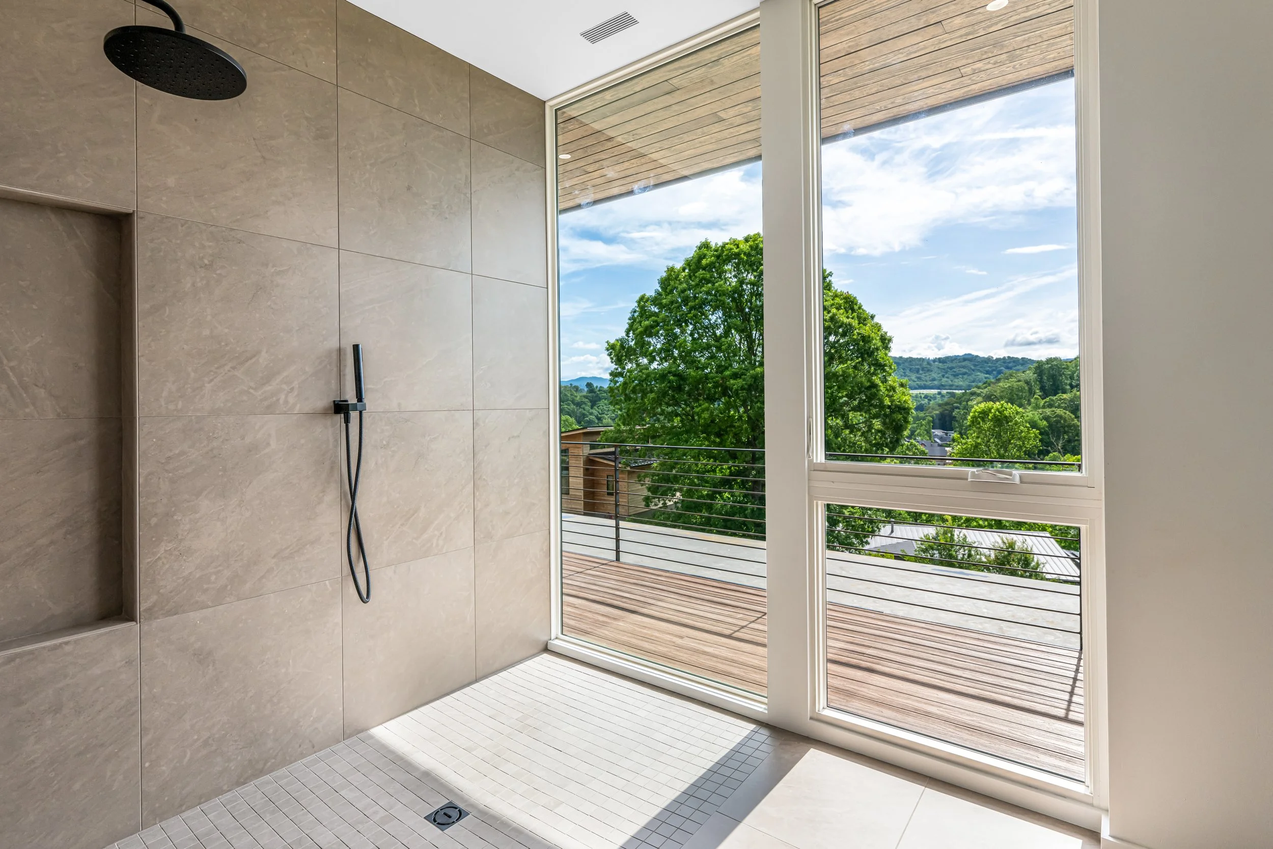 Modern shower area with beige tiles, black showerhead and handheld shower, large glass window showing green trees and blue sky outside, wooden balcony railing, bright natural light.