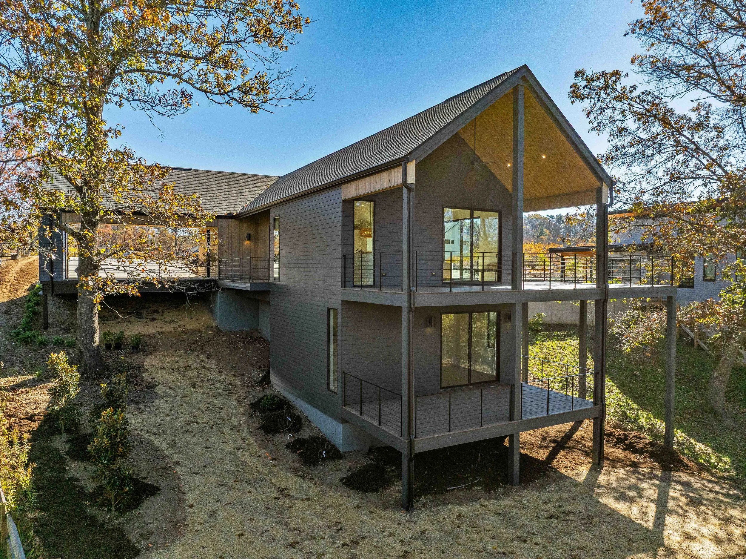 Modern multi-story house with large glass windows and wraparound balconies, situated on a hillside with trees and landscaping.