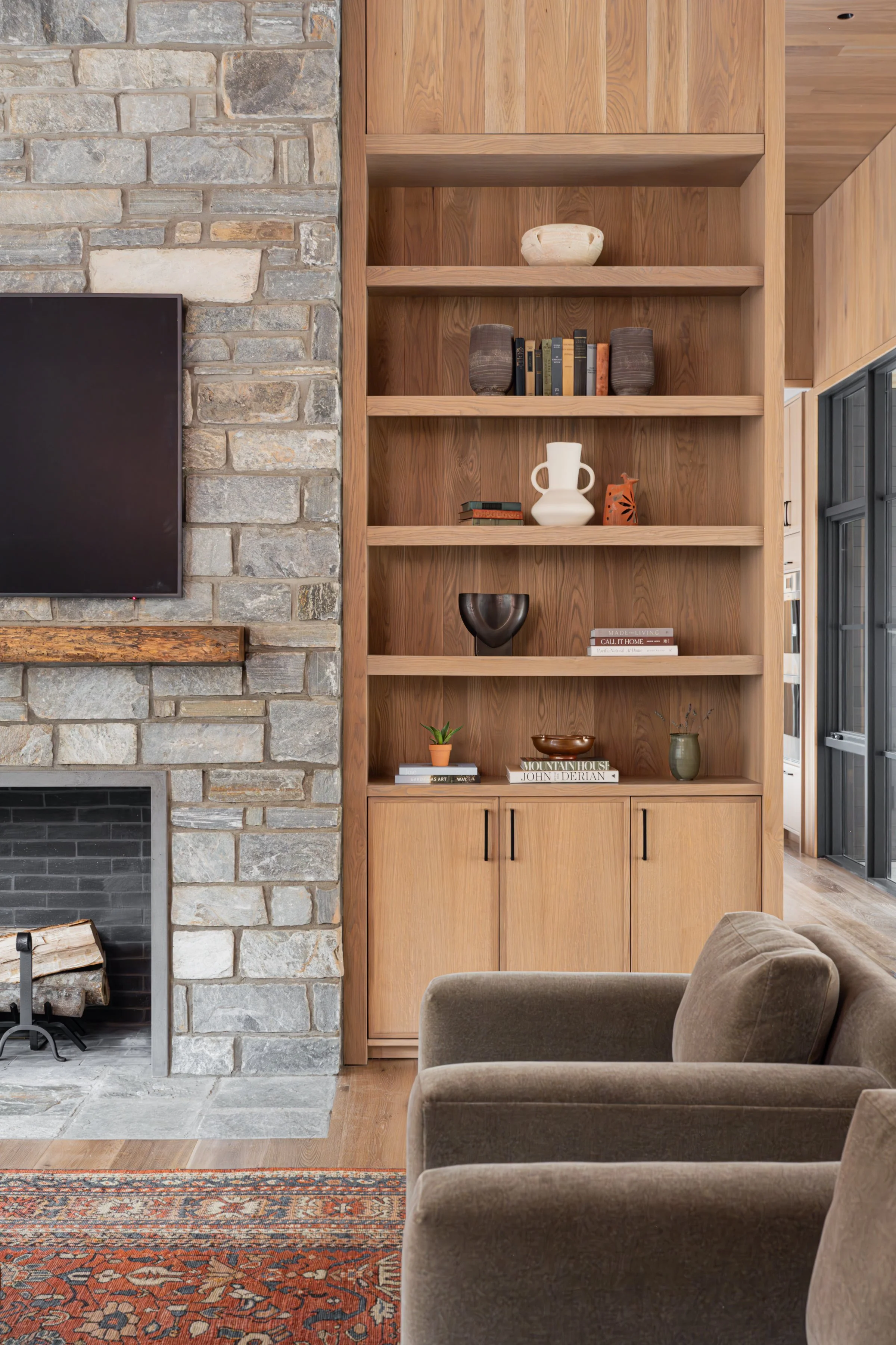 Interior of a living room with a stone and brick fireplace, a wooden shelf with decorative items, and part of a beige armchair.