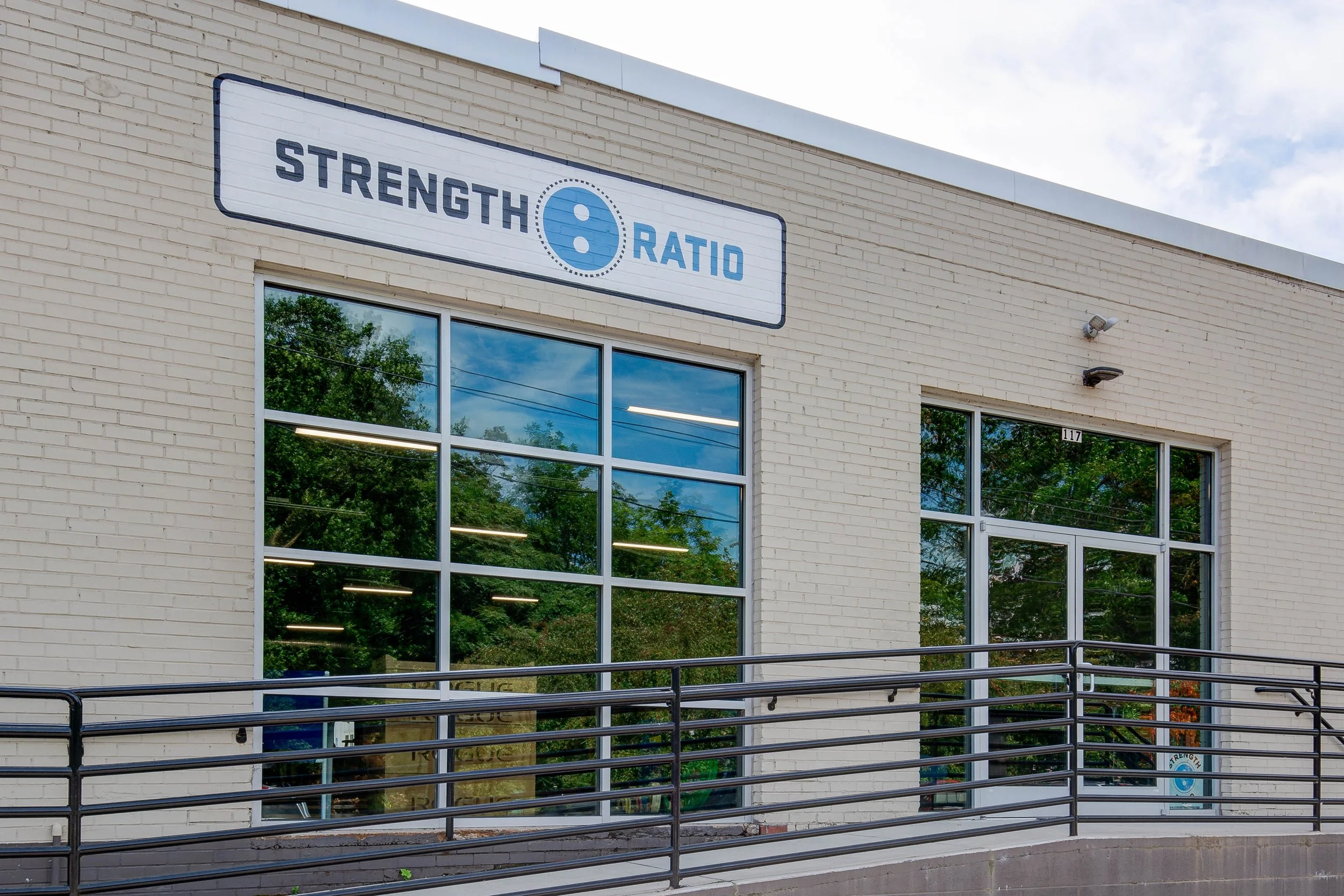 Exterior of a building with a sign reading 'Strength 8 Ratio' above a large window, ref lecting trees and a cloudy sky.