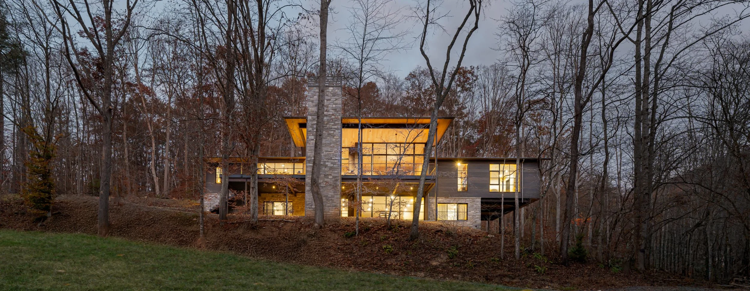 A modern house built on a hillside surrounded by leafless trees, with large glass windows and warm interior lighting visible at dusk.