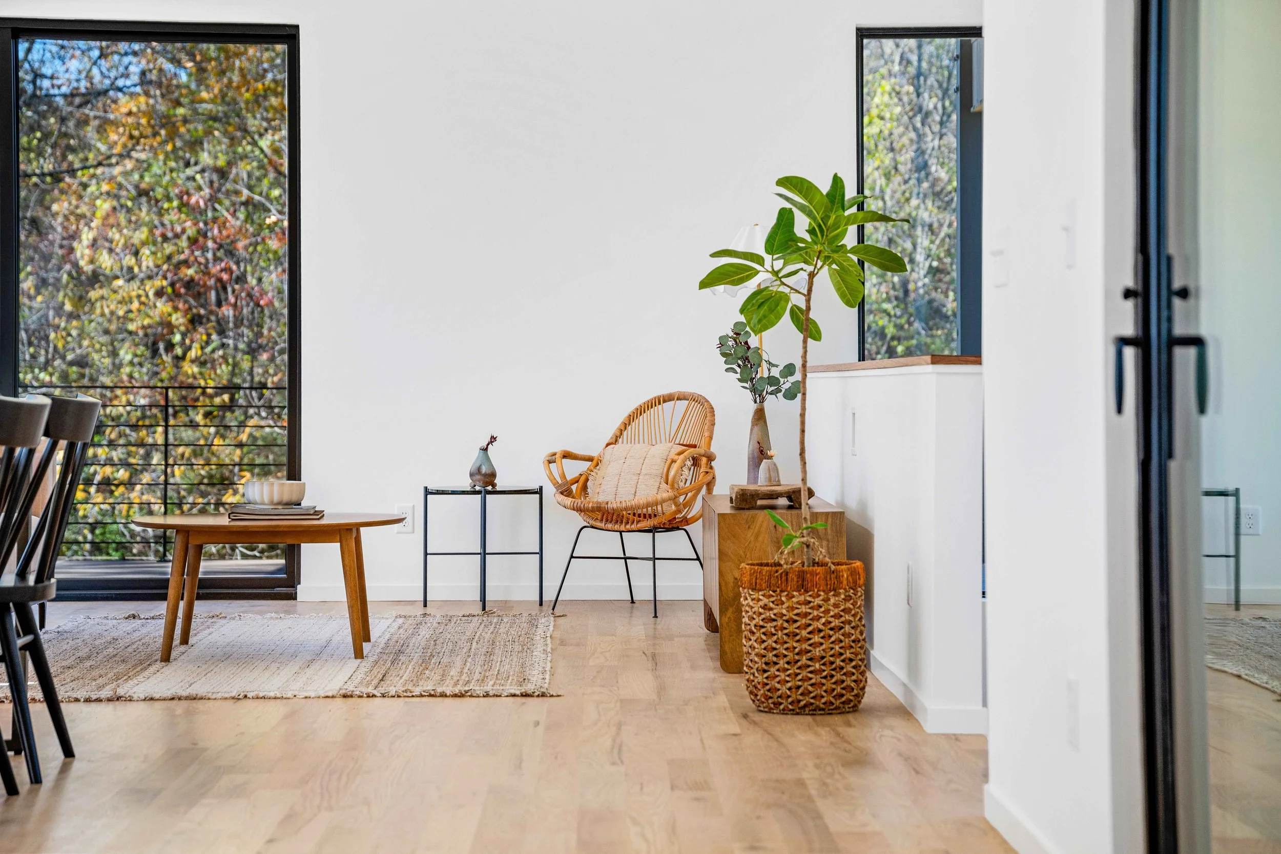 Minimalist living room with wood floors, large windows showing trees, a wicker chair, potted plant, small side tables, and a rug.