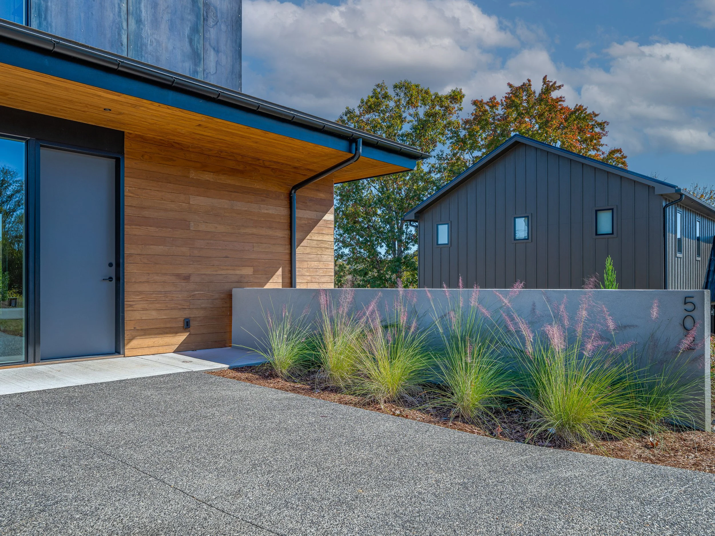 Modern house with wooden and metal exterior, concrete driveway, and landscaped plants in front, under partly cloudy sky.