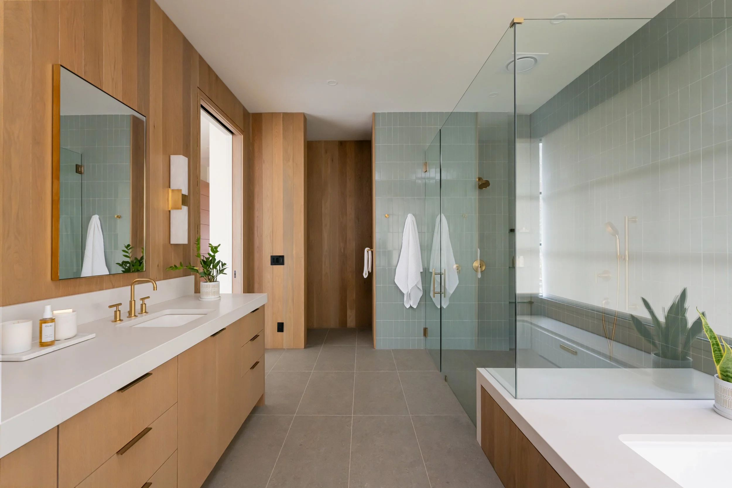 Modern bathroom with wooden cabinets, a large mirror, a double sink with gold fixtures, a walk-in shower with glass walls, white towels, and green plants.