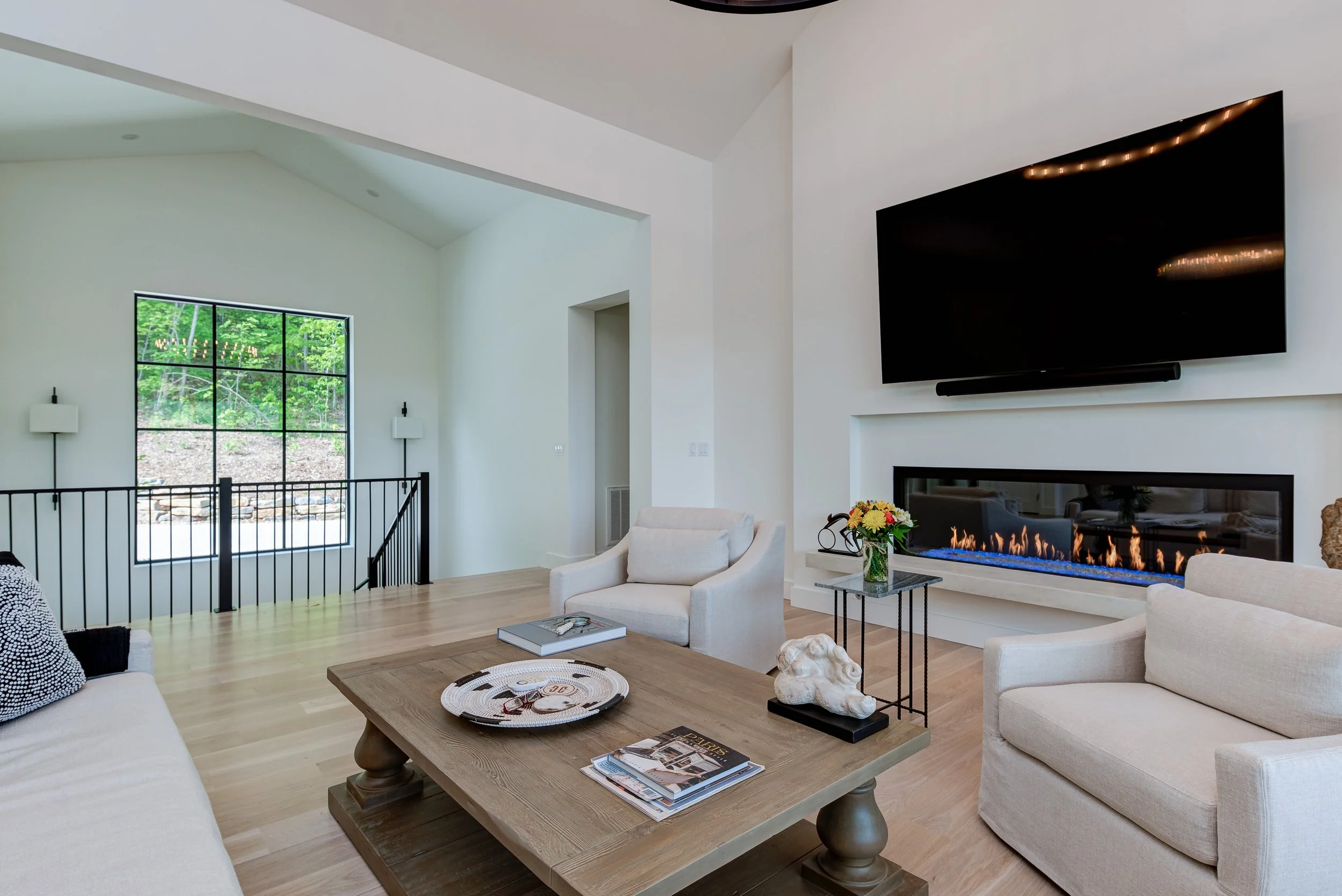 Living room with white walls, a fireplace beneath a large flat-screen TV, light-colored seating, a wooden coffee table with magazines and decorative items, a small side table with a flower vase, and a large window showing green trees outside.
