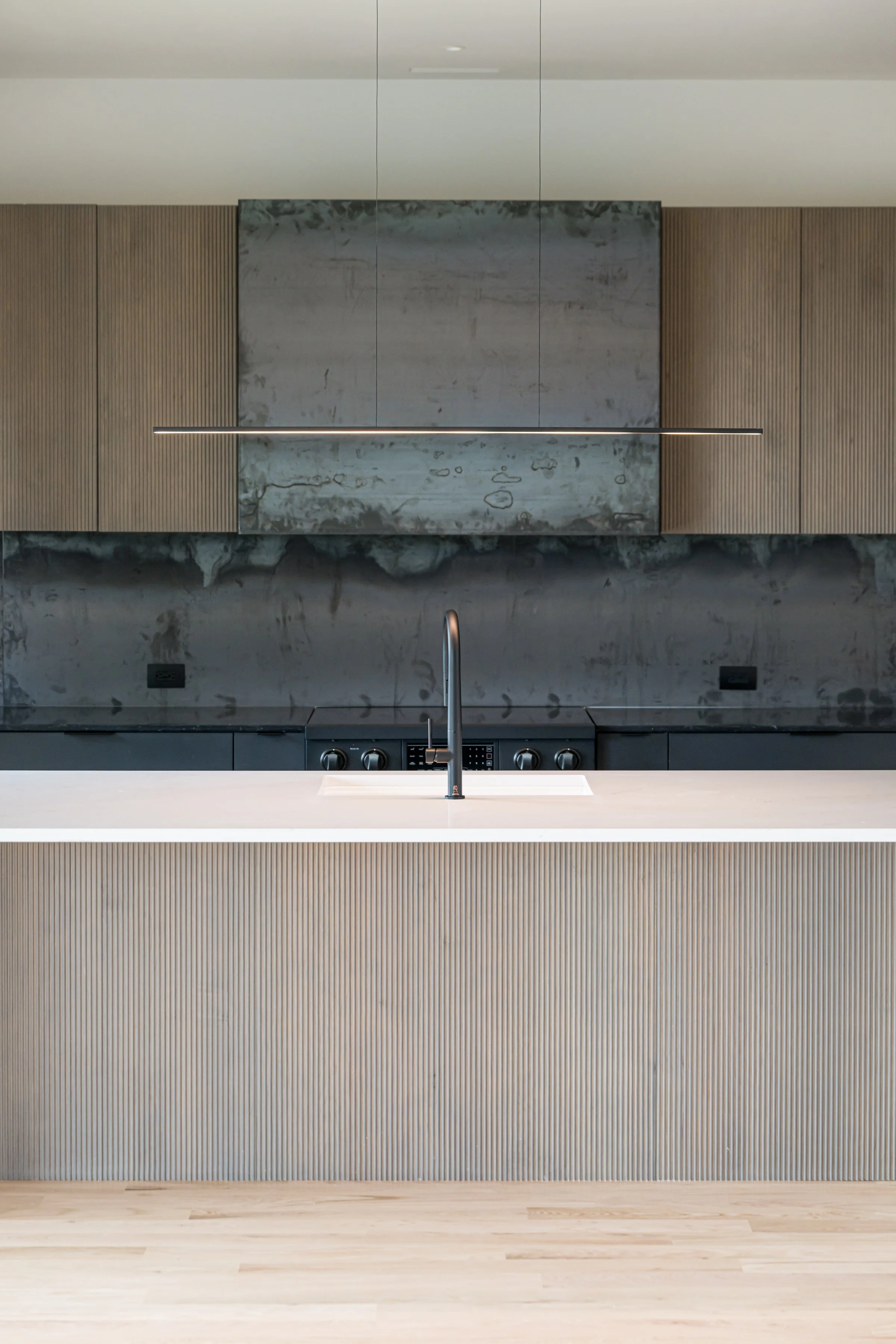 Modern kitchen with minimalist design, featuring a white countertop, a black faucet, and a sleek stovetop against a textured gray backsplash and wooden cabinets.