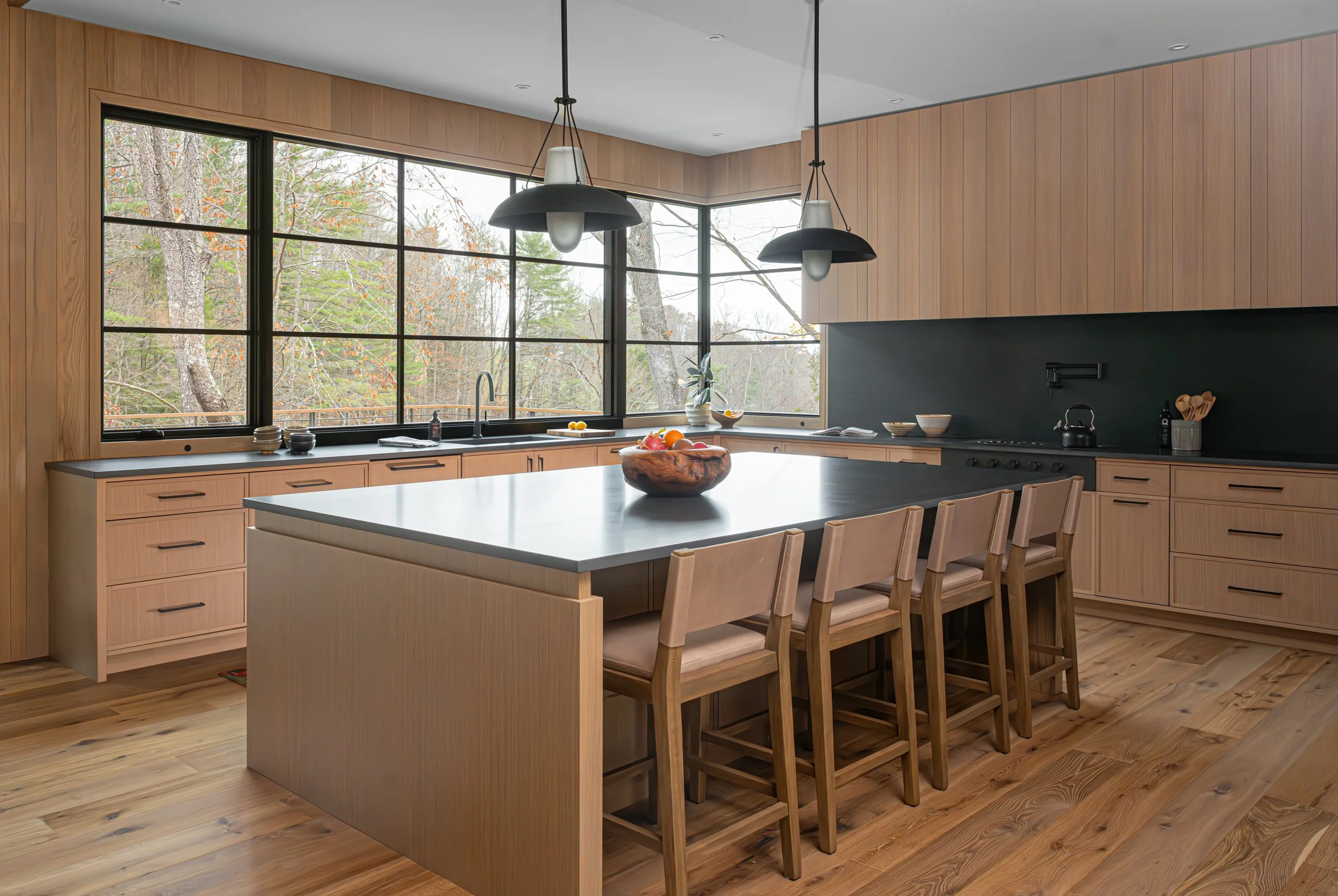 Modern kitchen with wooden cabinetry, large window views of trees, kitchen island with wooden chairs, black countertops, pendant lights, and minimal decor.