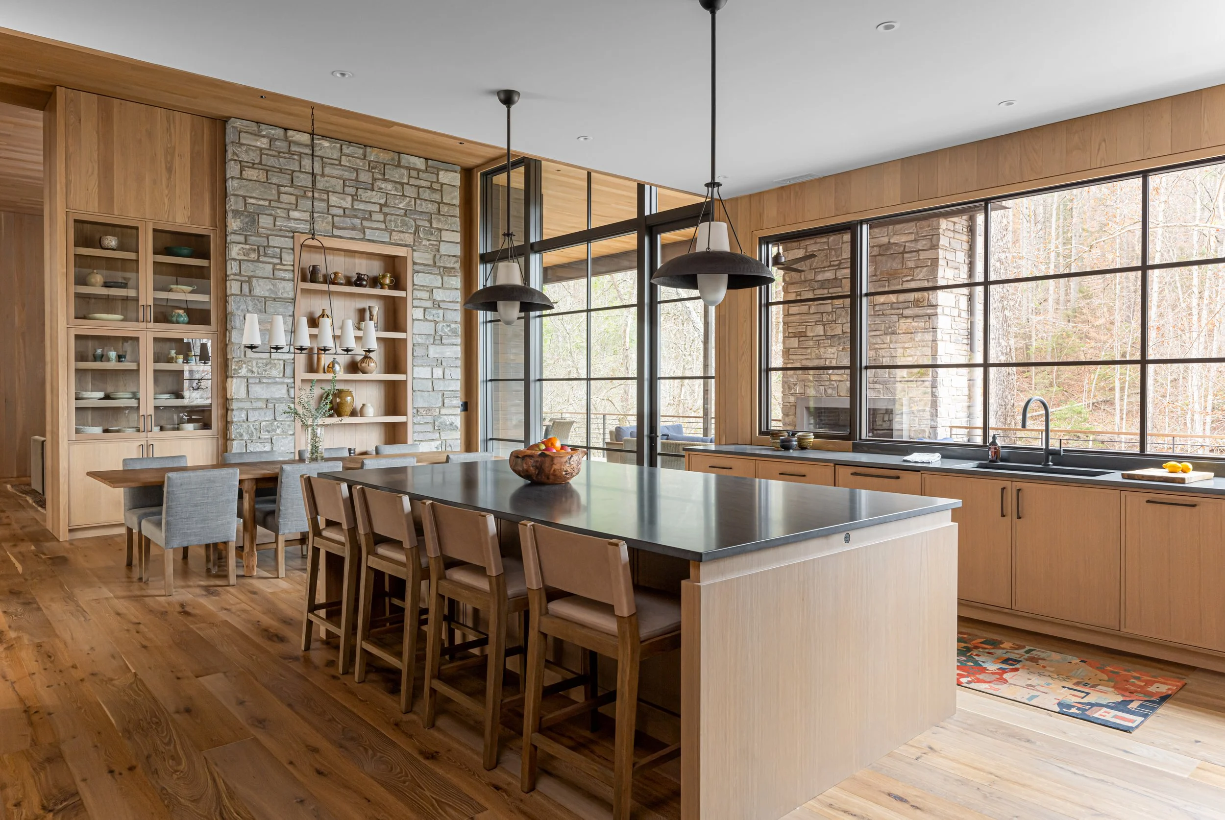 Modern kitchen with wooden cabinets, stone accent wall, large island with a dark countertop, pendant lighting, large windows with a view of trees, and hardwood flooring.