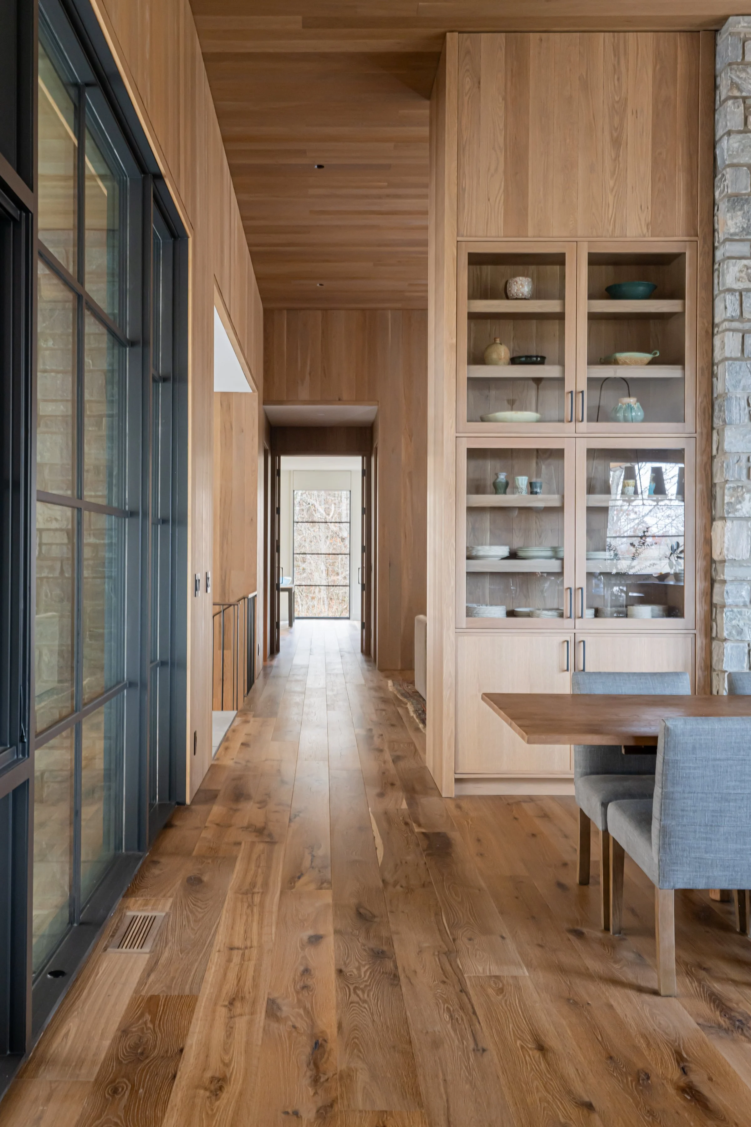 Interior view of a modern home with wooden walls, floors, and ceiling, a glass-fronted cabinet with dishes, gray upholstered chairs, and a hallway leading to a room with large windows.