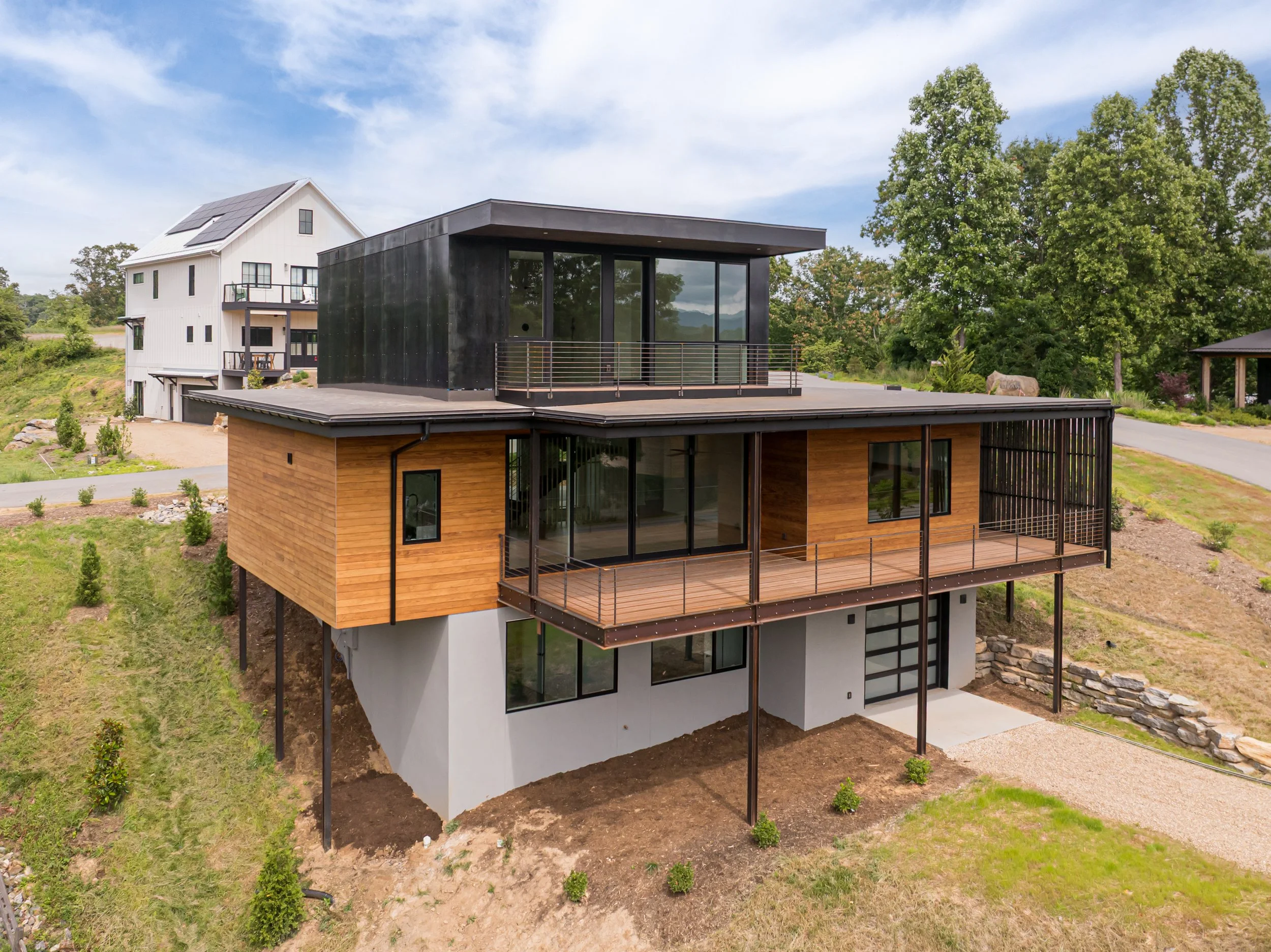Modern multi-story house on a hillside with large glass windows, wooden and black exterior finishes, and multiple balconies, surrounded by trees and a dirt yard.