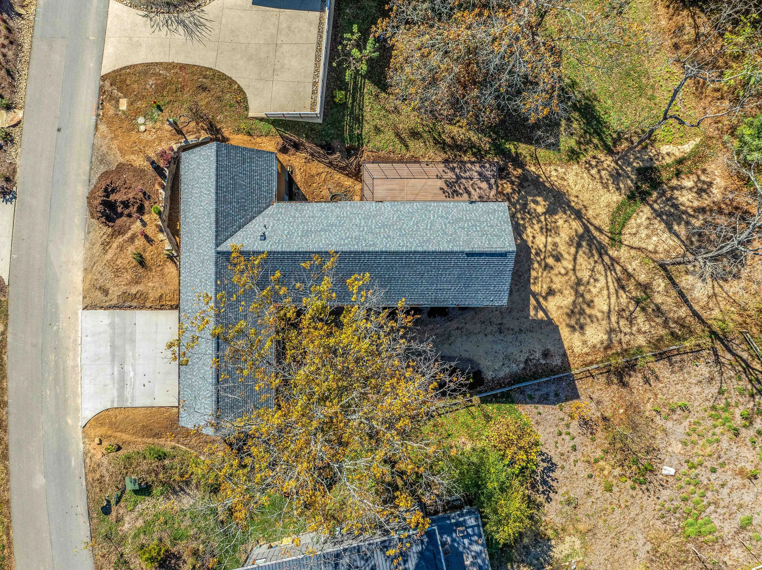 Aerial view of a house with a gray roof, surrounding trees, a front yard with a tree with yellow leaves, with leafless trees casting shadows, neighboring yard with a shed, and a street on the left side.