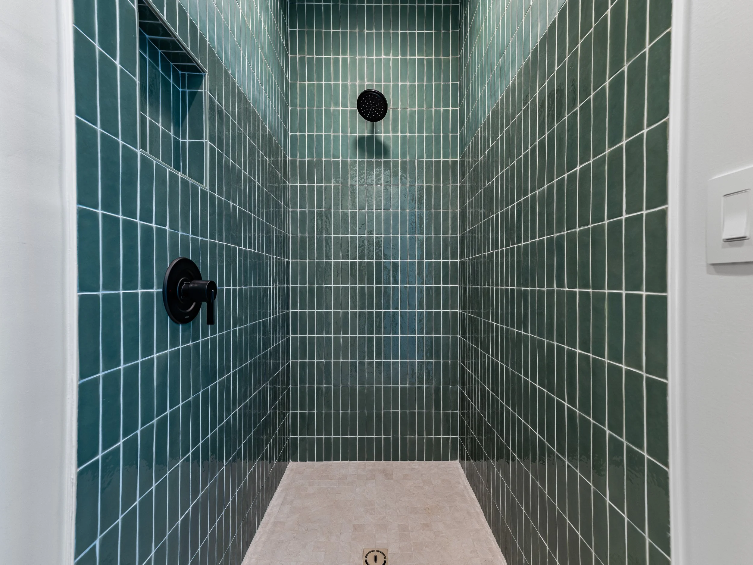 Empty green-tiled shower with a black showerhead, black handle, built-in shelf, and white walls.