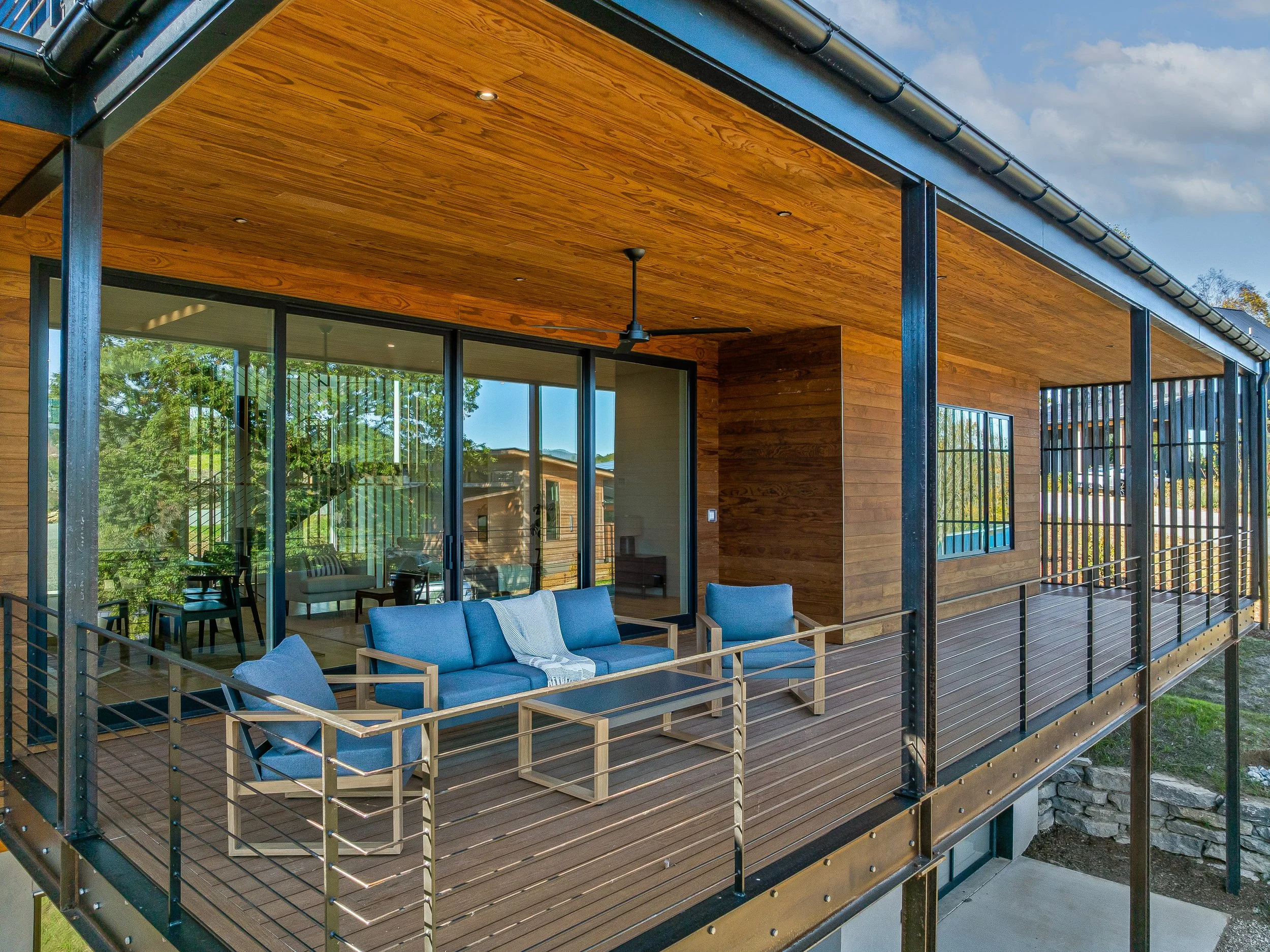 Modern house with a large balcony featuring a wooden ceiling and black railing, furnished with blue chairs and a table, with glass sliding doors and windows reflecting trees and a neighboring house.