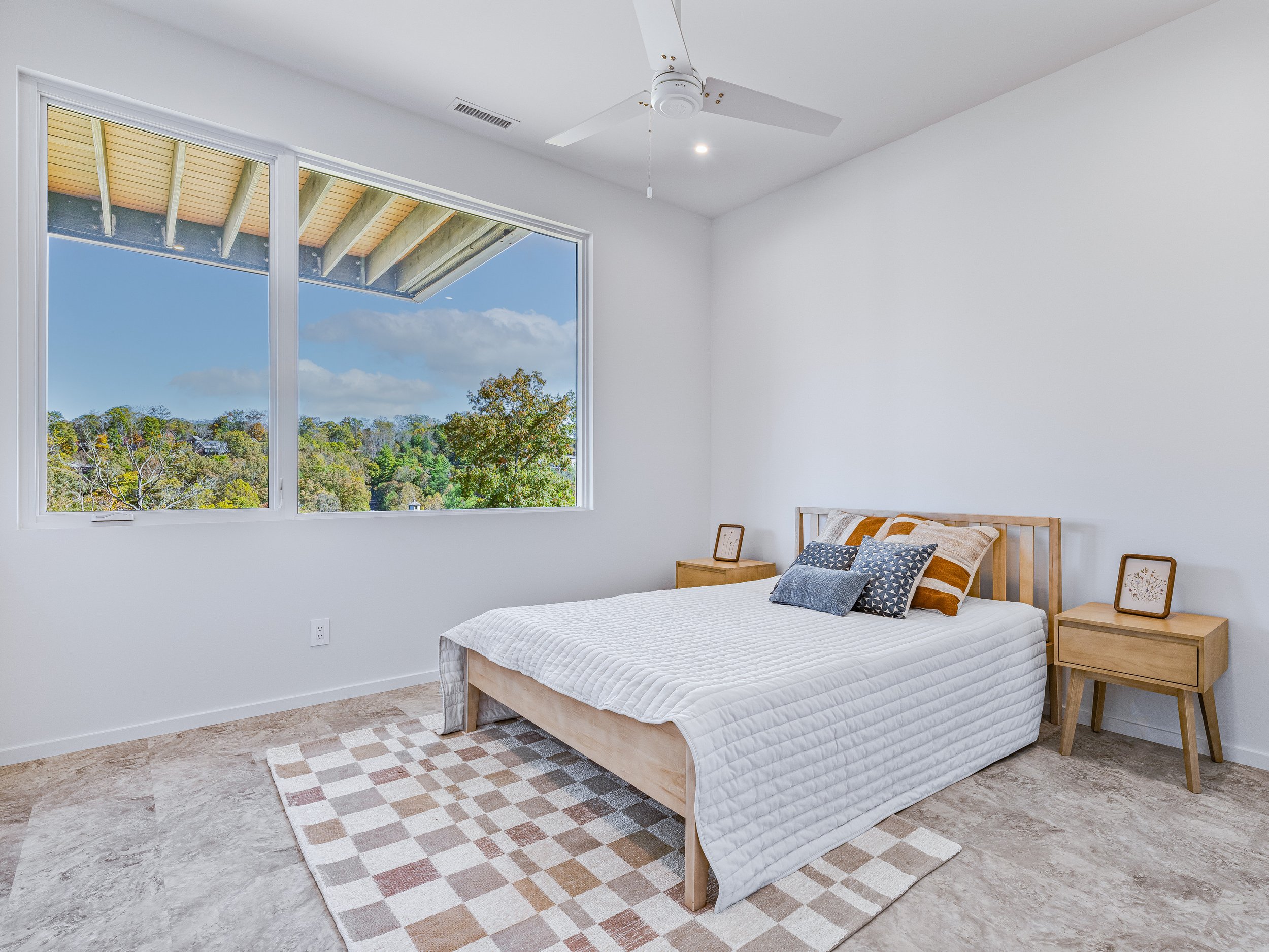 A bright bedroom with a large window showing trees and a blue sky outside. The room has a wooden bed frame with a white quilt, three pillows, and two wooden nightstands with framed pictures. There is a ceiling fan and a patterned rug on the beige til