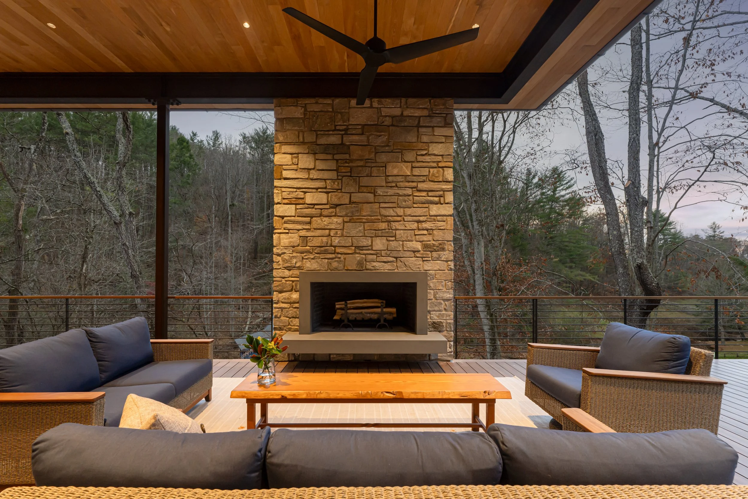 Outdoor patio with a stone fireplace, wooden ceiling, ceiling fan, and surrounded by trees, featuring sofas, a wooden table, and a vase with flowers.