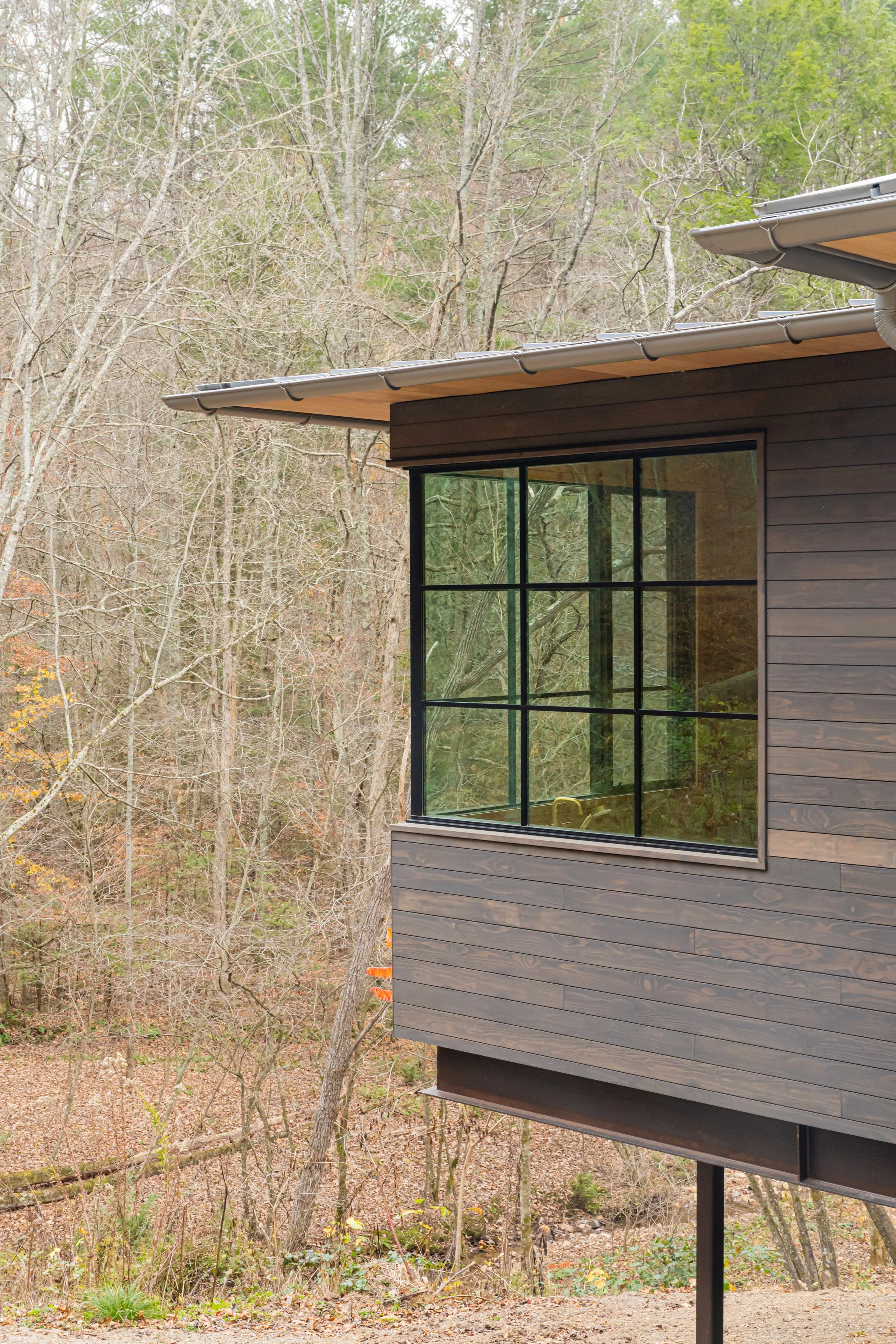 Corner of modern house with large glass window, wooden siding, and sloped roof in a wooded area.