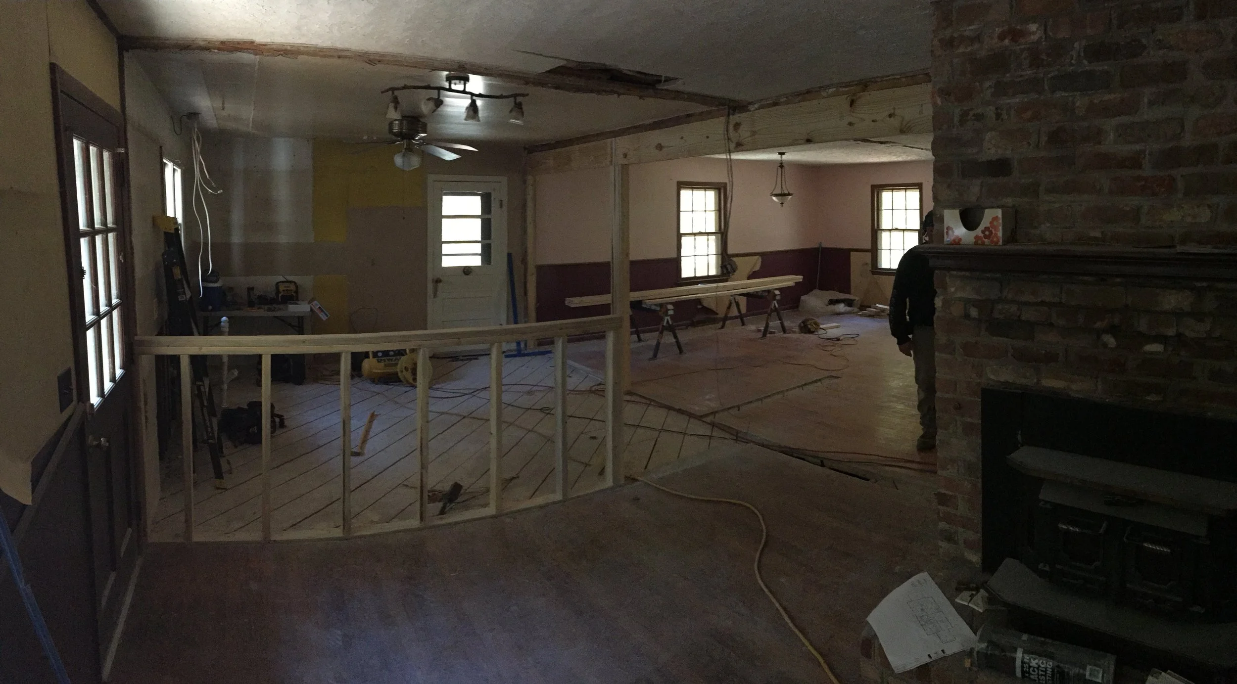 Interior of a house under renovation with partially installed flooring, construction tools, a ceiling fan, and a man working near a brick fireplace.