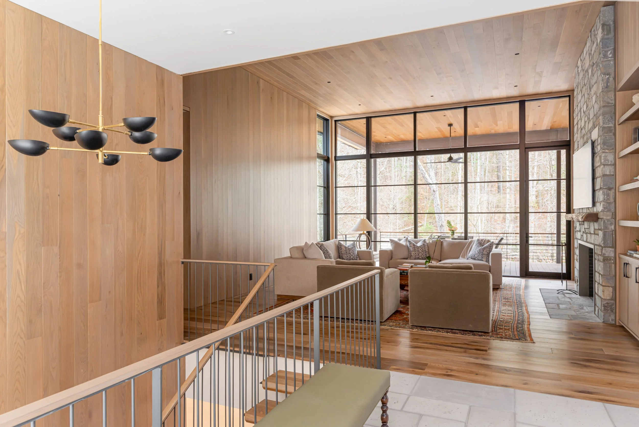 Living room with large window, beige sofas, wooden wall, stone fireplace, wooden shelves, and a modern black sputnik chandelier.