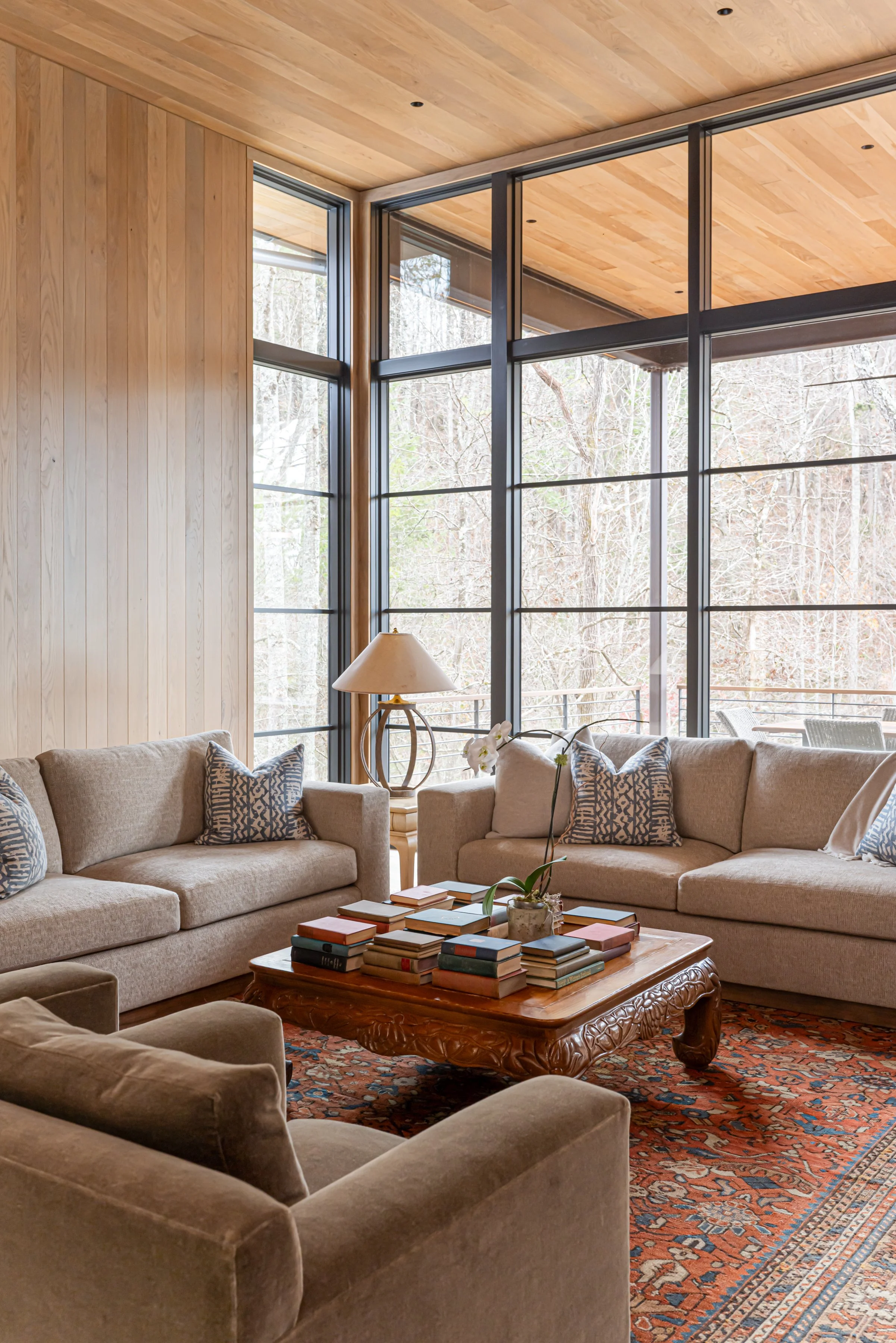 Living room with large glass windows, beige sofas with patterned pillows, a wooden coffee table with stacked books, a lamp, and a patterned rug.