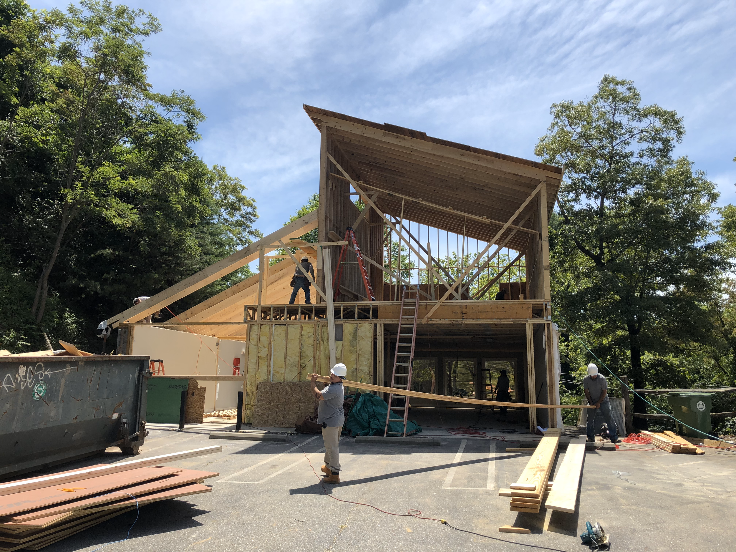Workers are constructing a wooden building in an outdoor area with trees and a partly cloudy sky.