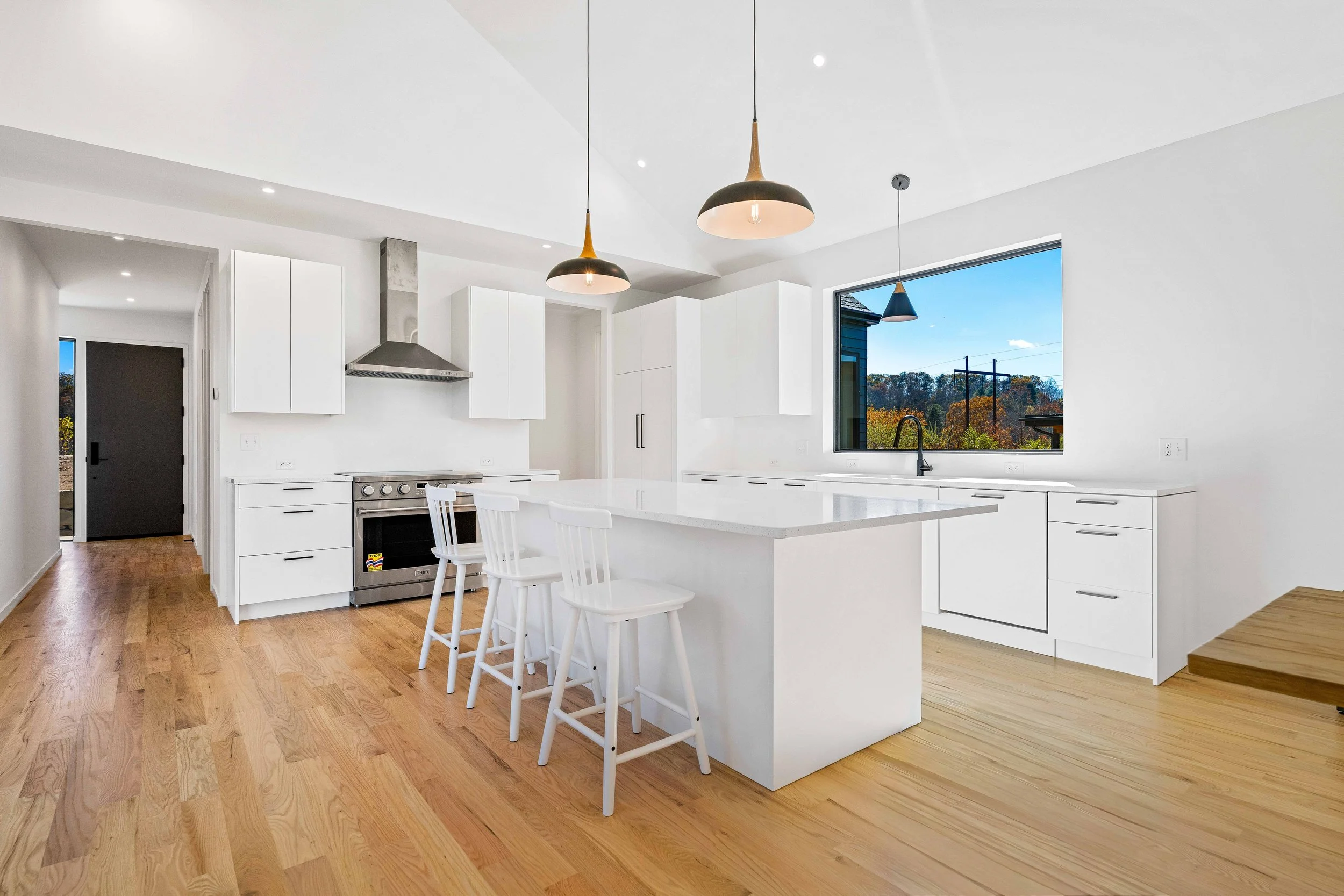 Modern white kitchen with wooden floor, island with three white chairs, large window with outdoor view, white cabinets, stainless steel stove, range hood, and pendant lights.