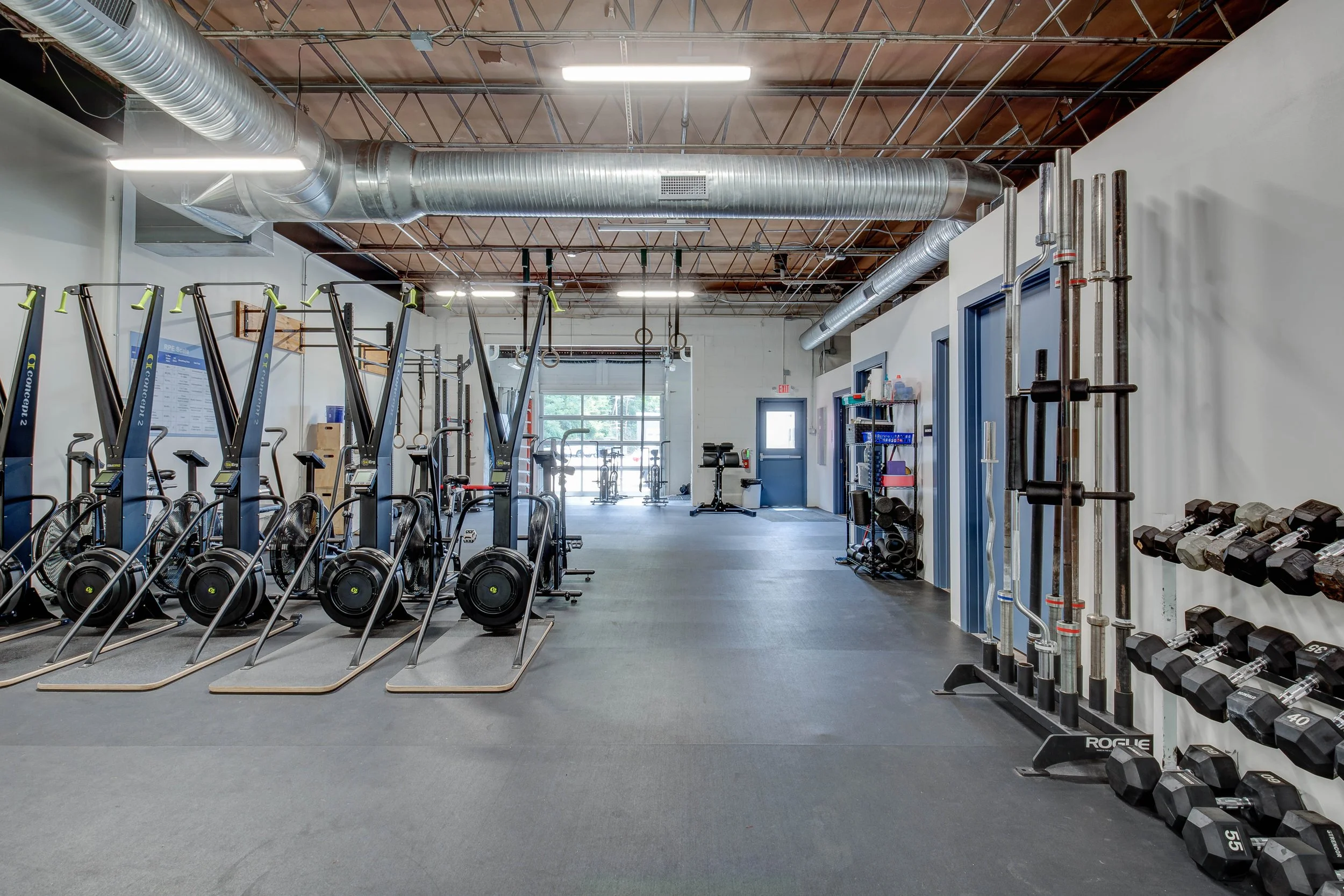 Fitness gym with rowers, free weights, and exercise equipment in a spacious room with high ceiling and exposed ductwork.
