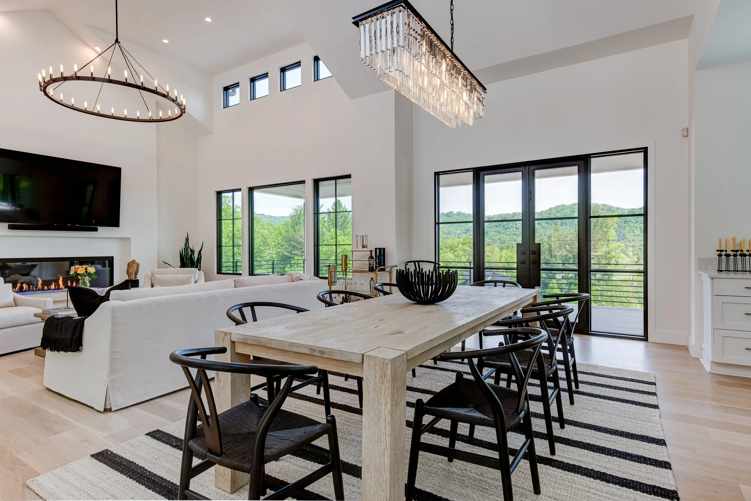 Modern living room with a dining area, large windows showing green landscape, black and white striped rug, white sofas, fireplace, and black pendant lighting.