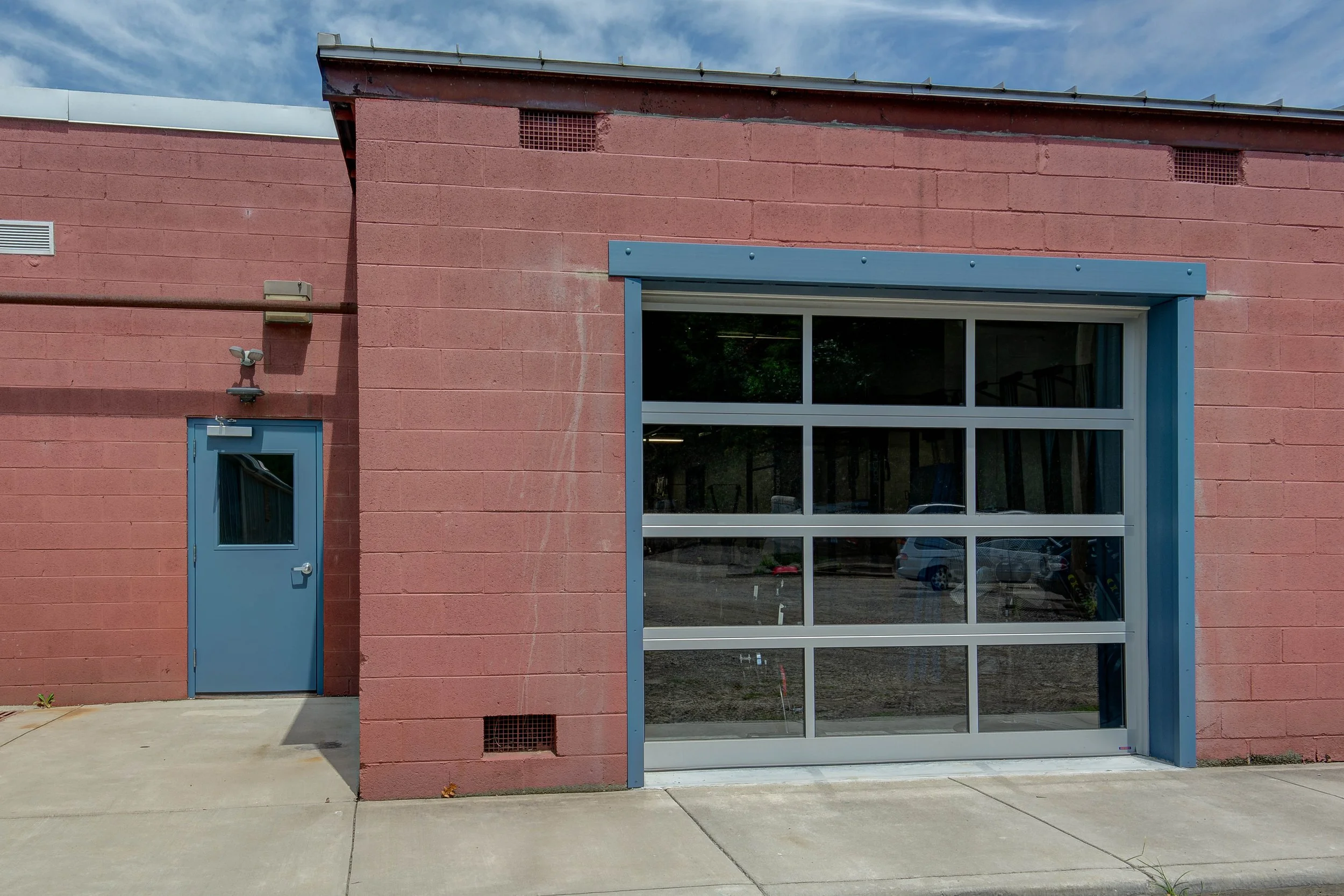 A red brick building with a blue metal door and a large glass roll-up garage door. The building has small vents near the roofline and on the lower wall, and is situated on a concrete sidewalk under a partly cloudy sky.