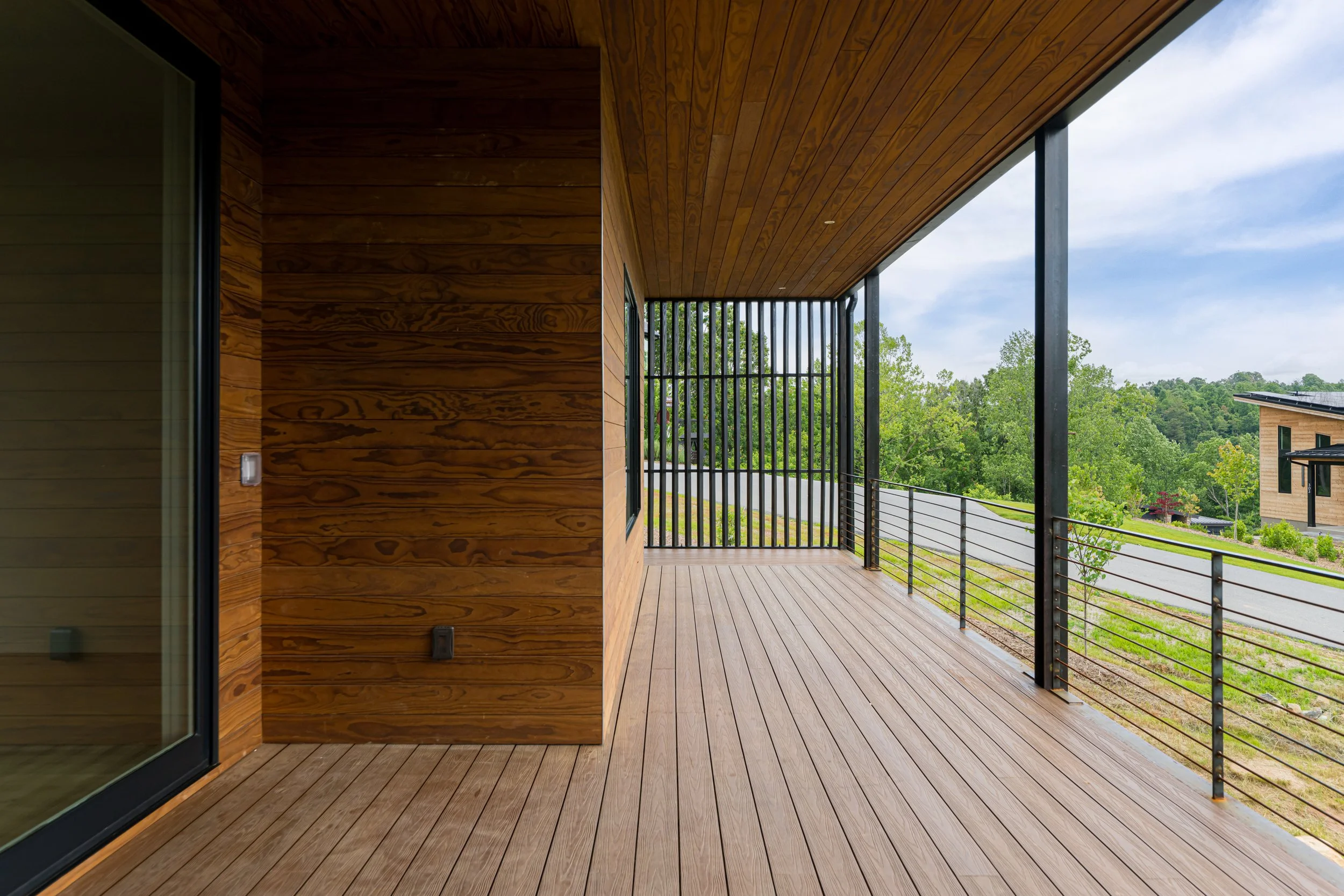 Wooden balcony with black metal railing, view of green trees and neighboring modern houses.