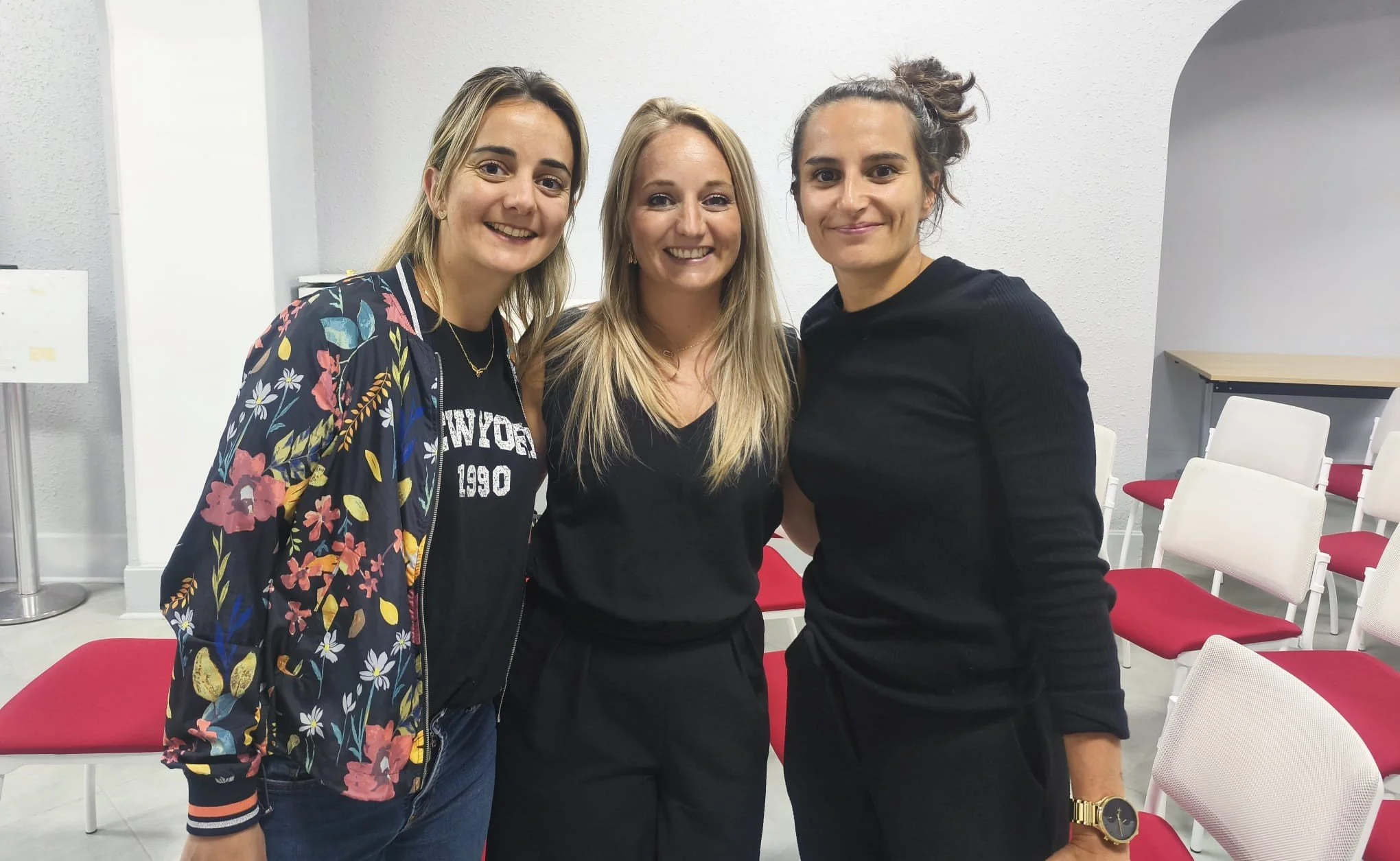 Trois femmes souriantes posent ensemble dans une salle, avec des chaises rouges et blanches en arrière-plan.