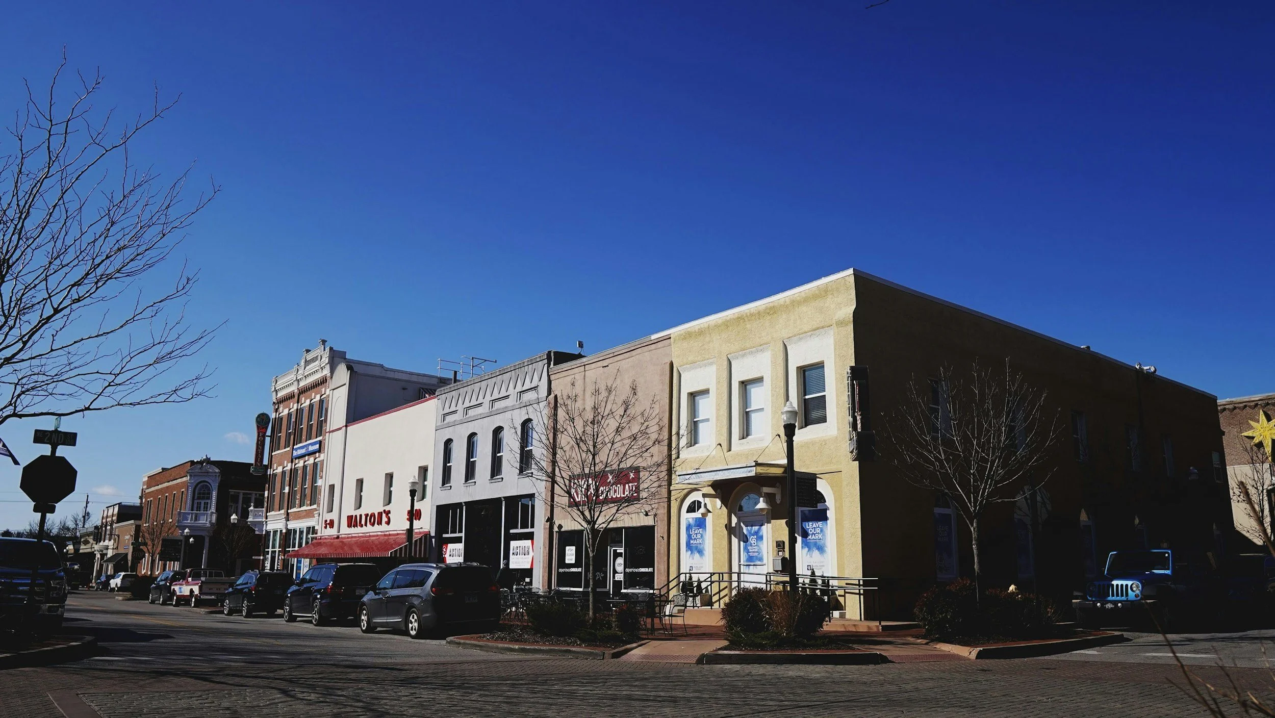 A small-town street with a row of colorful storefronts, parked cars along the curb, leafless trees, and a clear blue sky.
