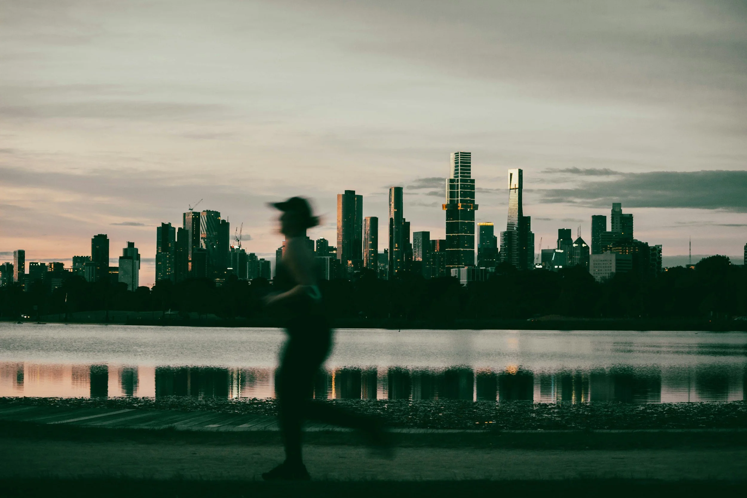 Silhouette of a person walking along a waterfront with a city skyline in the background during dusk.