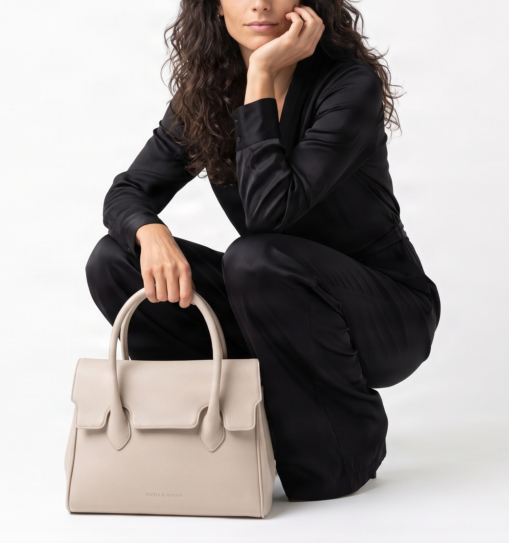 A woman with wavy brown hair, dressed in black satin pajamas, crouching next to a beige handbag with a flap closure and a handle, against a plain white background.