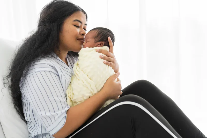 Woman holding and snuggling a newborn baby wrapped in a yellow blanket, sitting in a bright room.